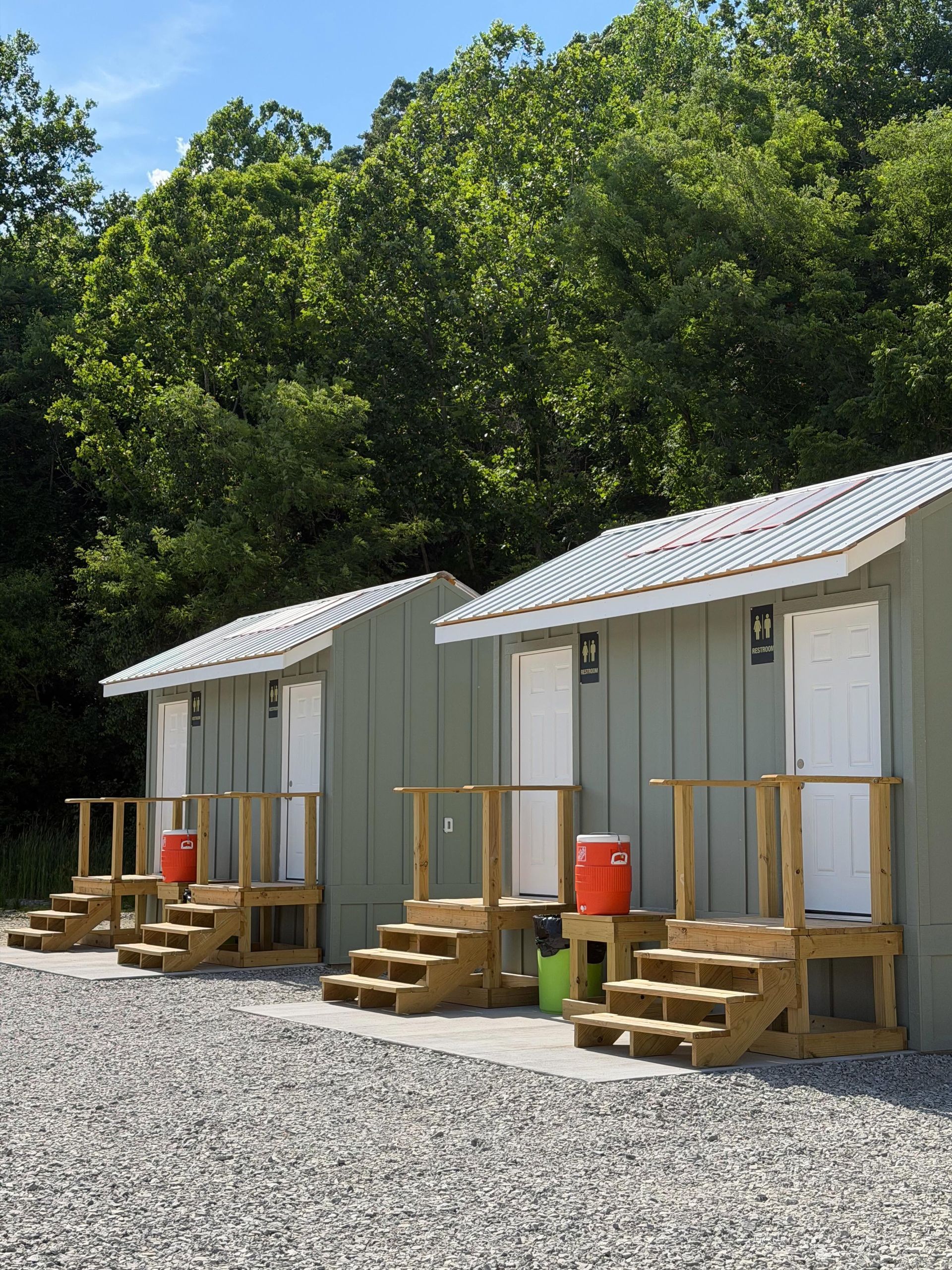 Two small white cabins with porches on gravel beside trees under a clear sky