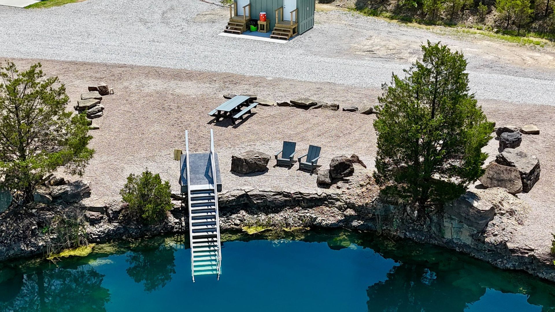 Aerial view of a lakeside dock with stairs, patio chairs, picnic table, and trees beside clear blue water