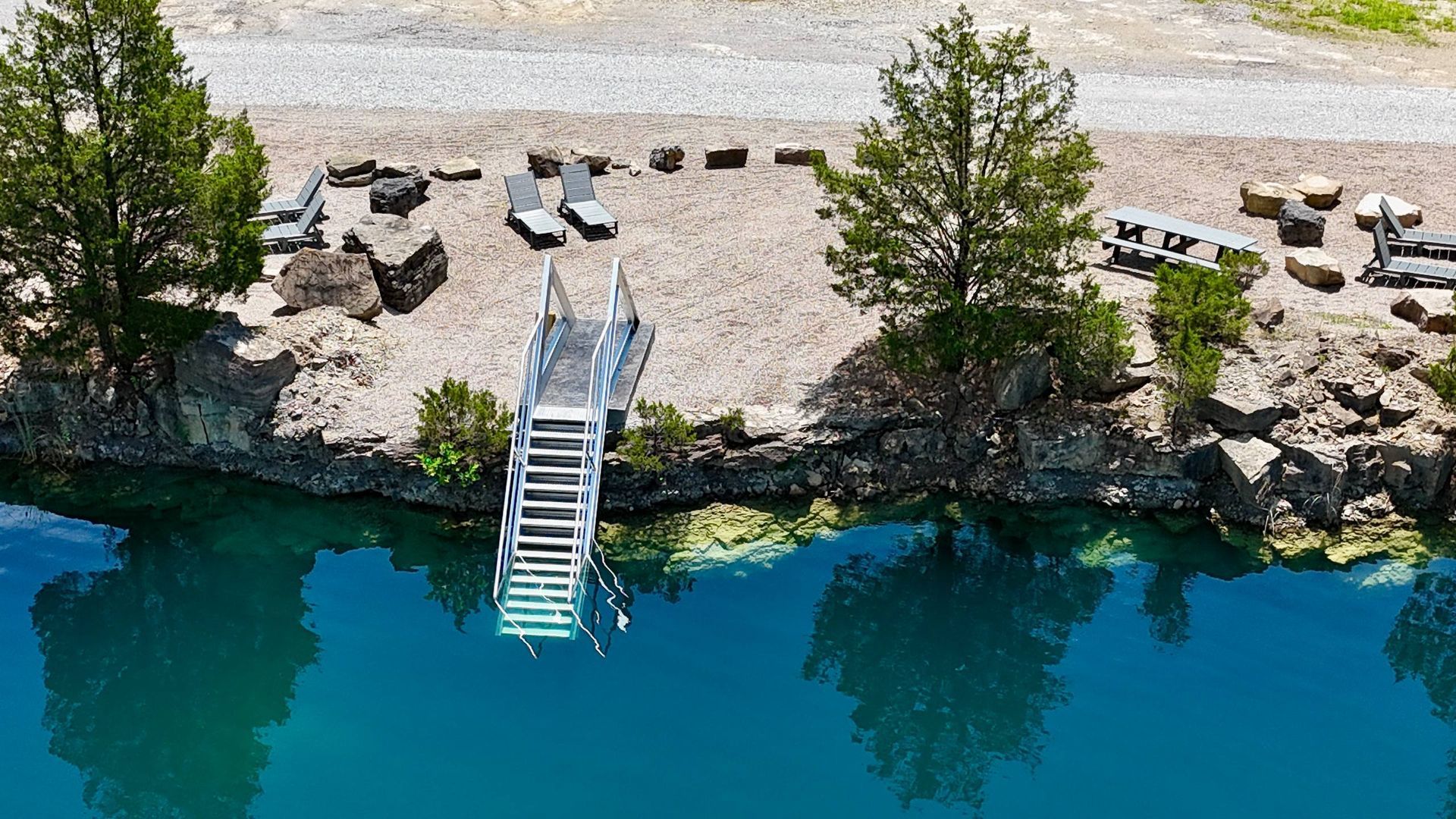 Lakeside dock with metal steps and a sandy shore lined with rocks and lounge chairs, beside clear blue water