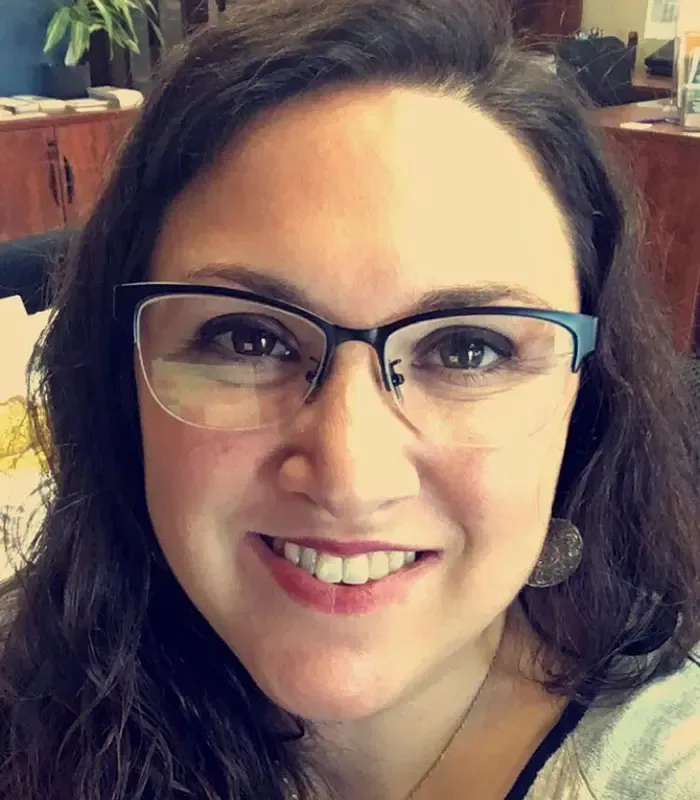 Woman with glasses smiles at the camera; dark hair, light skin, office setting.