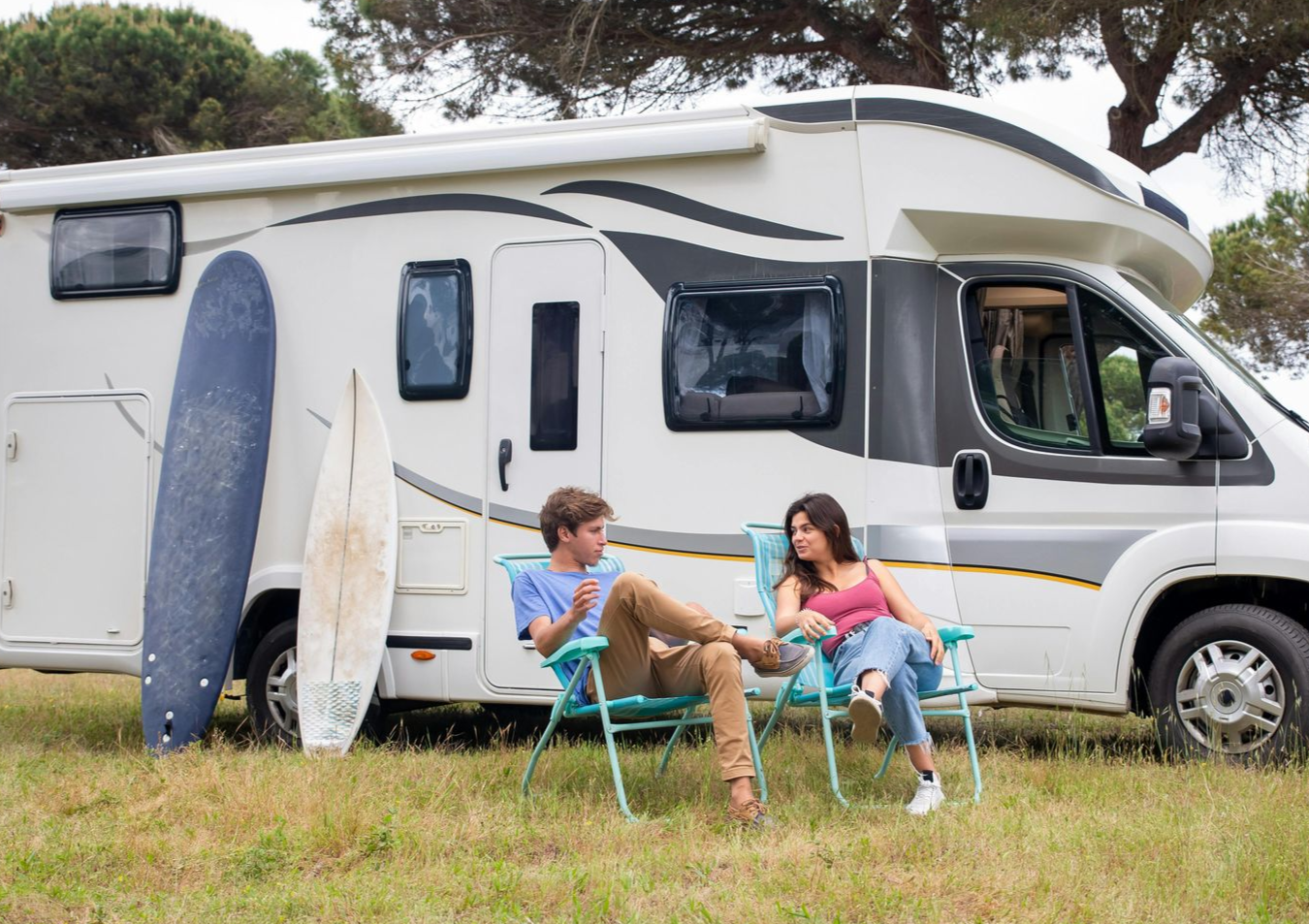 Couple in chairs near RV in field, surfboard leans against it.