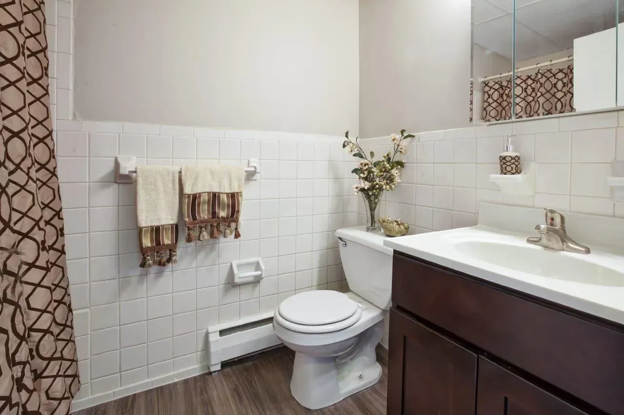 Bright apartment bathroom with white tile walls, dark wood vanity, toilet, and patterned shower curtain.