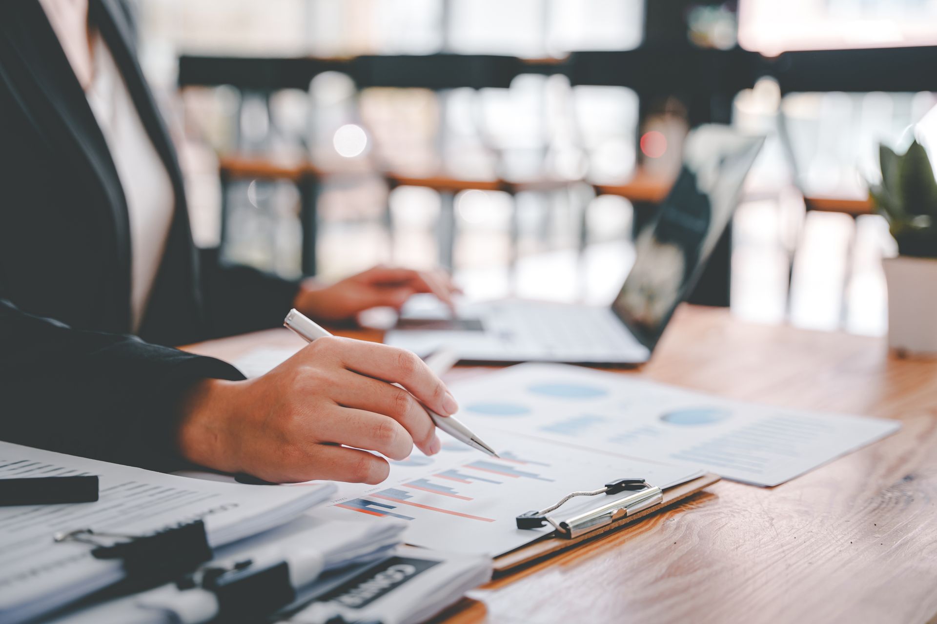 Hand holding a pen over charts on a clipboard at a desk with a laptop and documents. Hand holding a pen over charts on a clipboard at a desk with a laptop and documents.