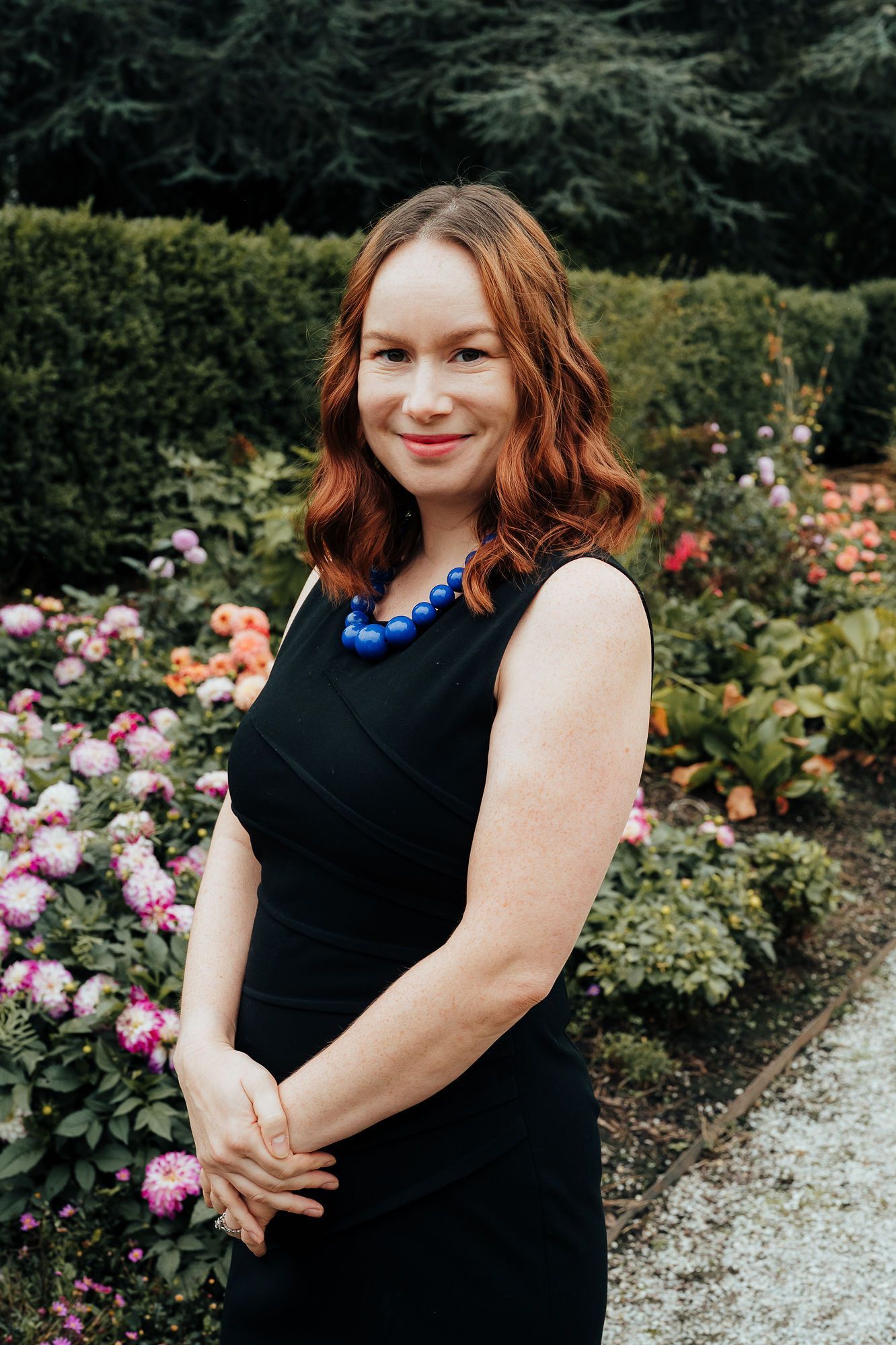 Woman in black dress, smiles at camera, standing in a flower garden.