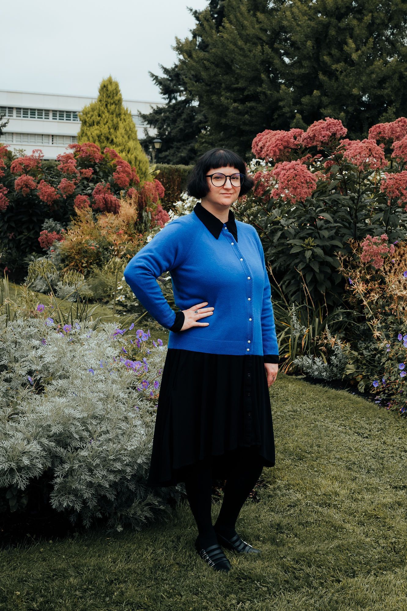 Woman in blue sweater and black skirt, poses in a garden.