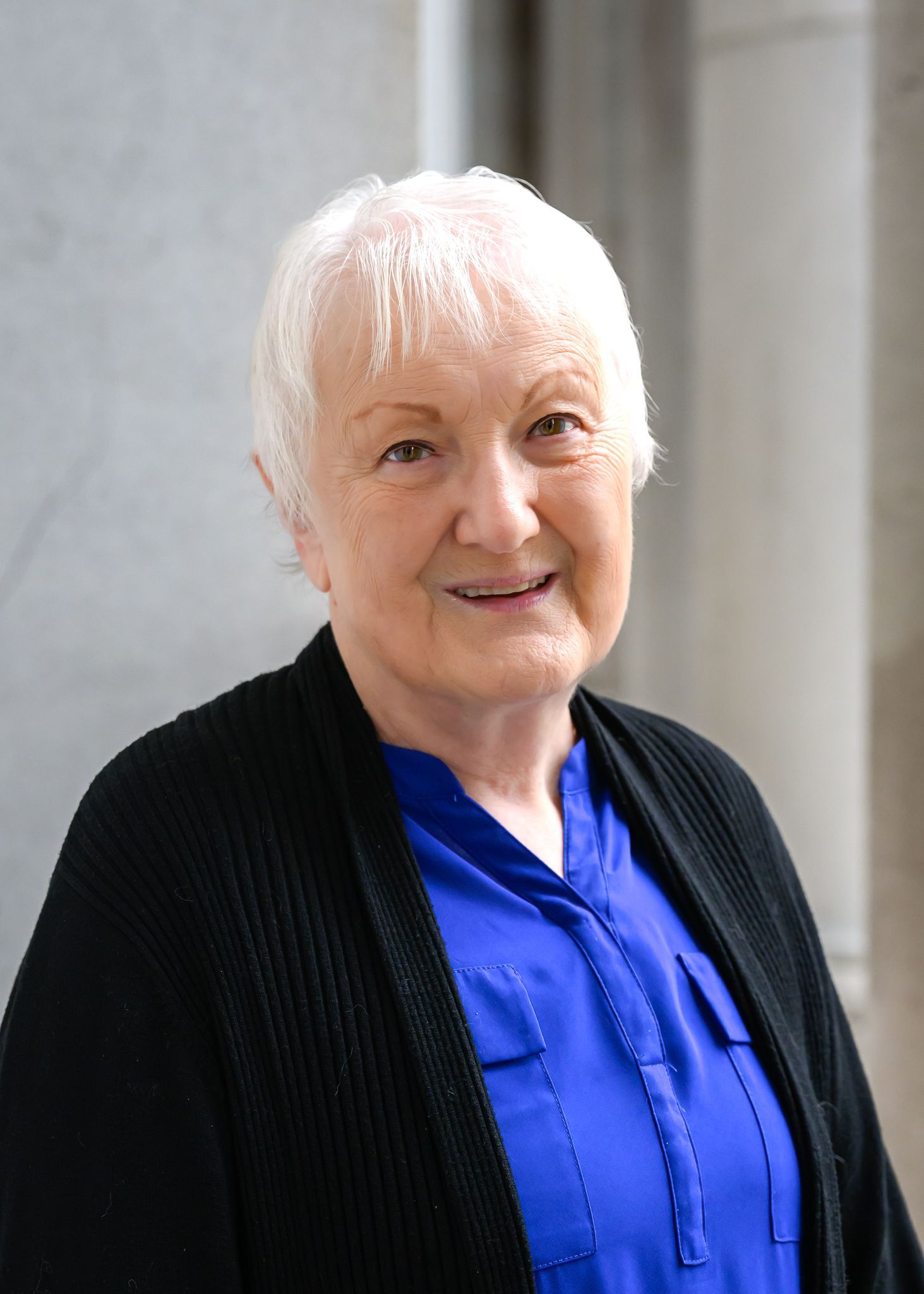 Woman with white hair in a blue shirt and black sweater smiles in a bright, neutral-toned setting.