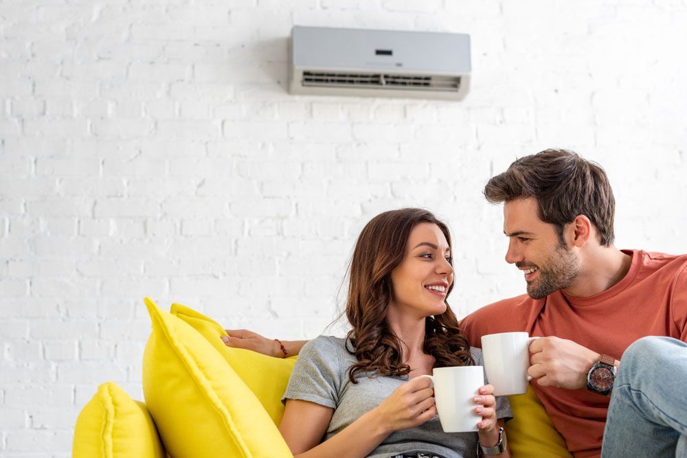 Couple smiling, holding mugs, sitting on yellow couch beneath a wall-mounted air conditioner.