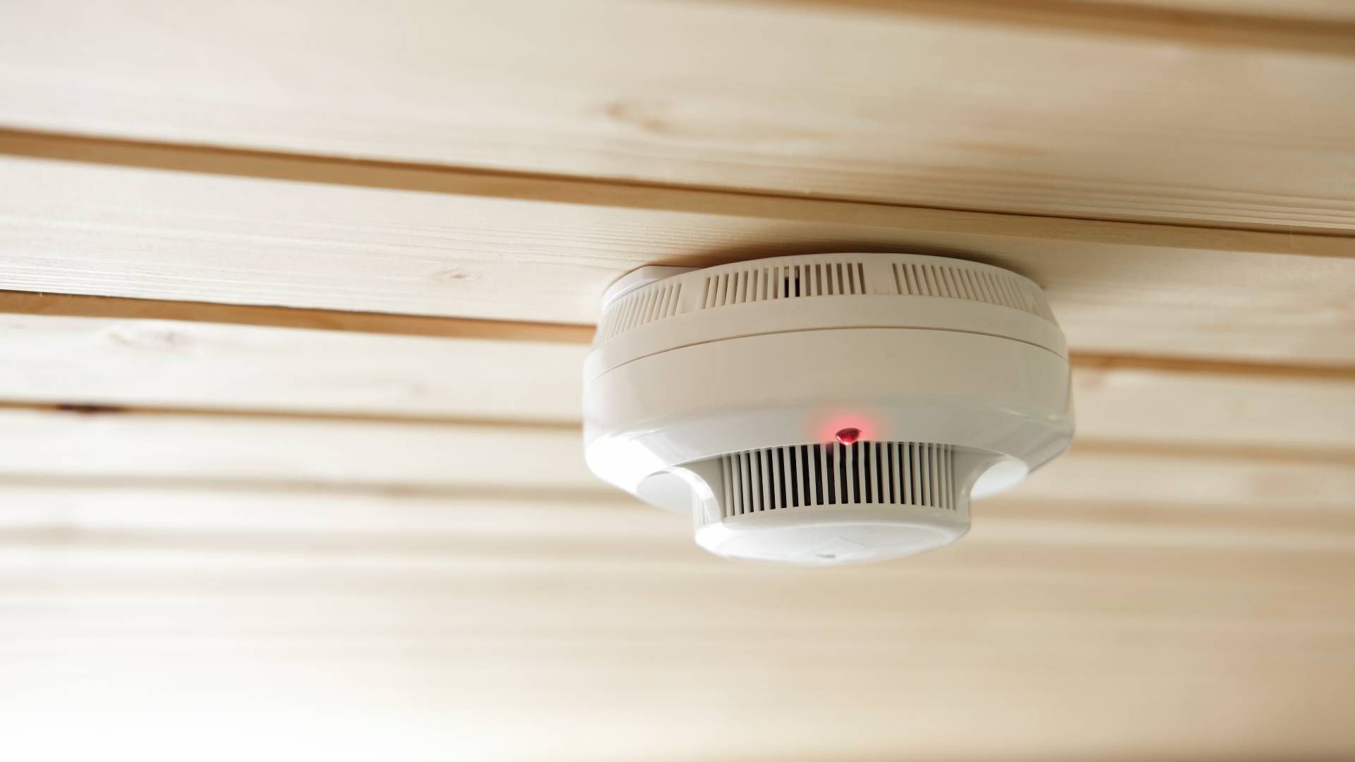 White smoke detector mounted on a beige ceiling, with a red indicator light.
