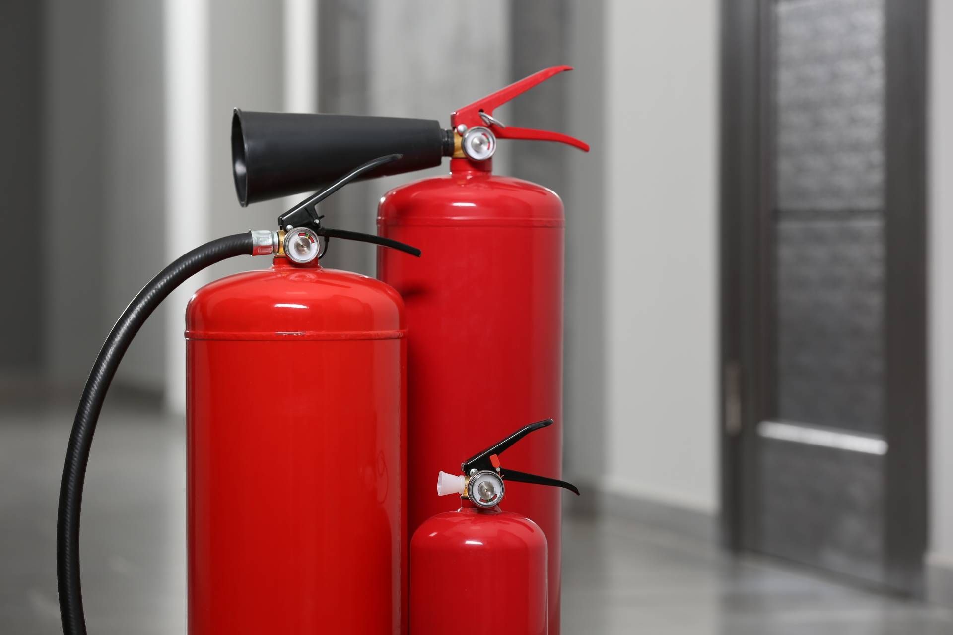 Three red fire extinguishers standing in a hallway, one with a black hose and one with a red horn.