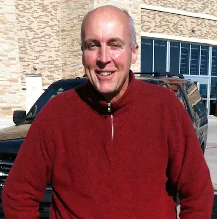 Man with short hair smiles in front of a building and black vehicle, wearing a red sweater.