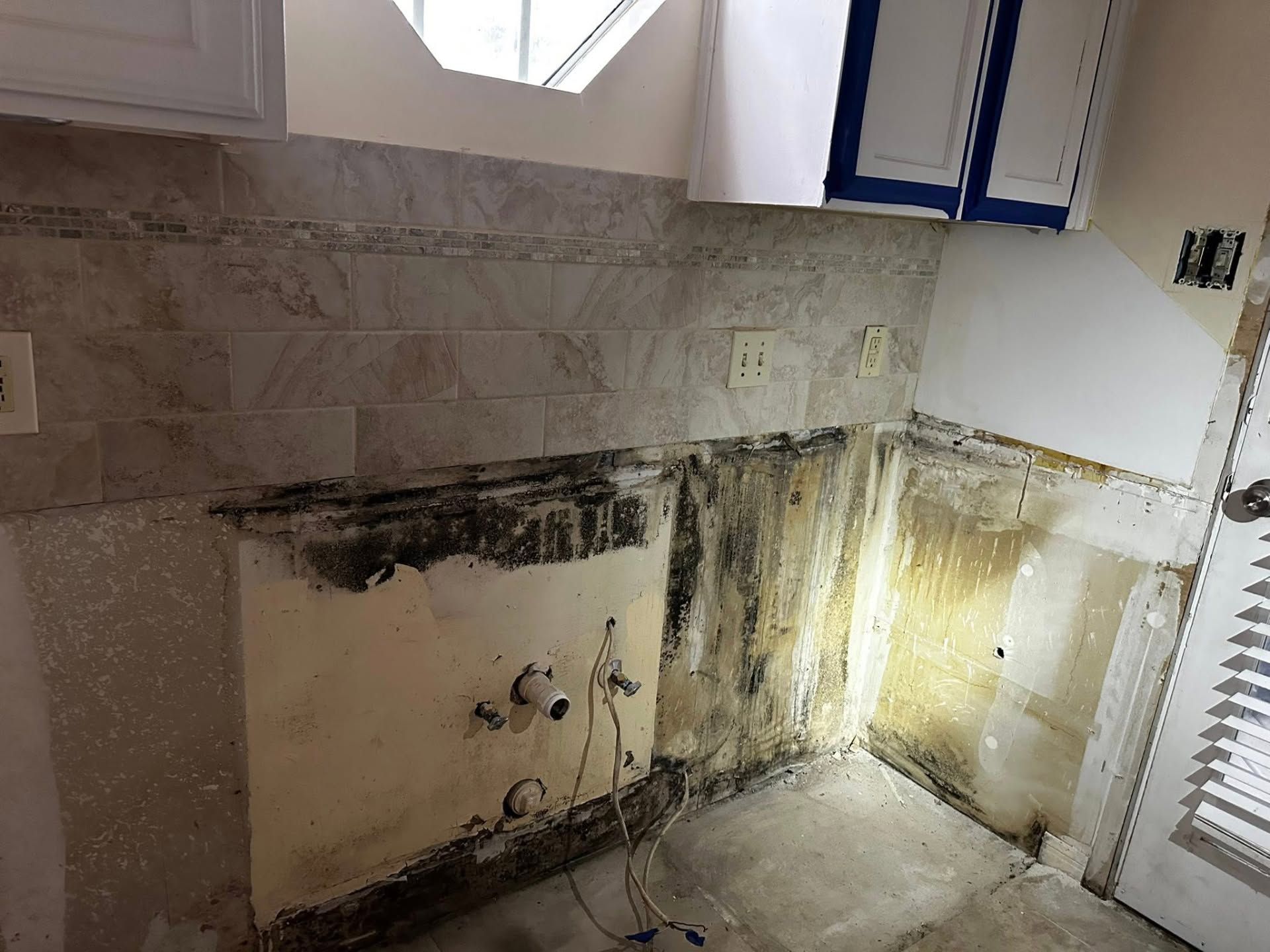 Damaged kitchen wall with mold stains, exposed wiring, and a window above a sink area