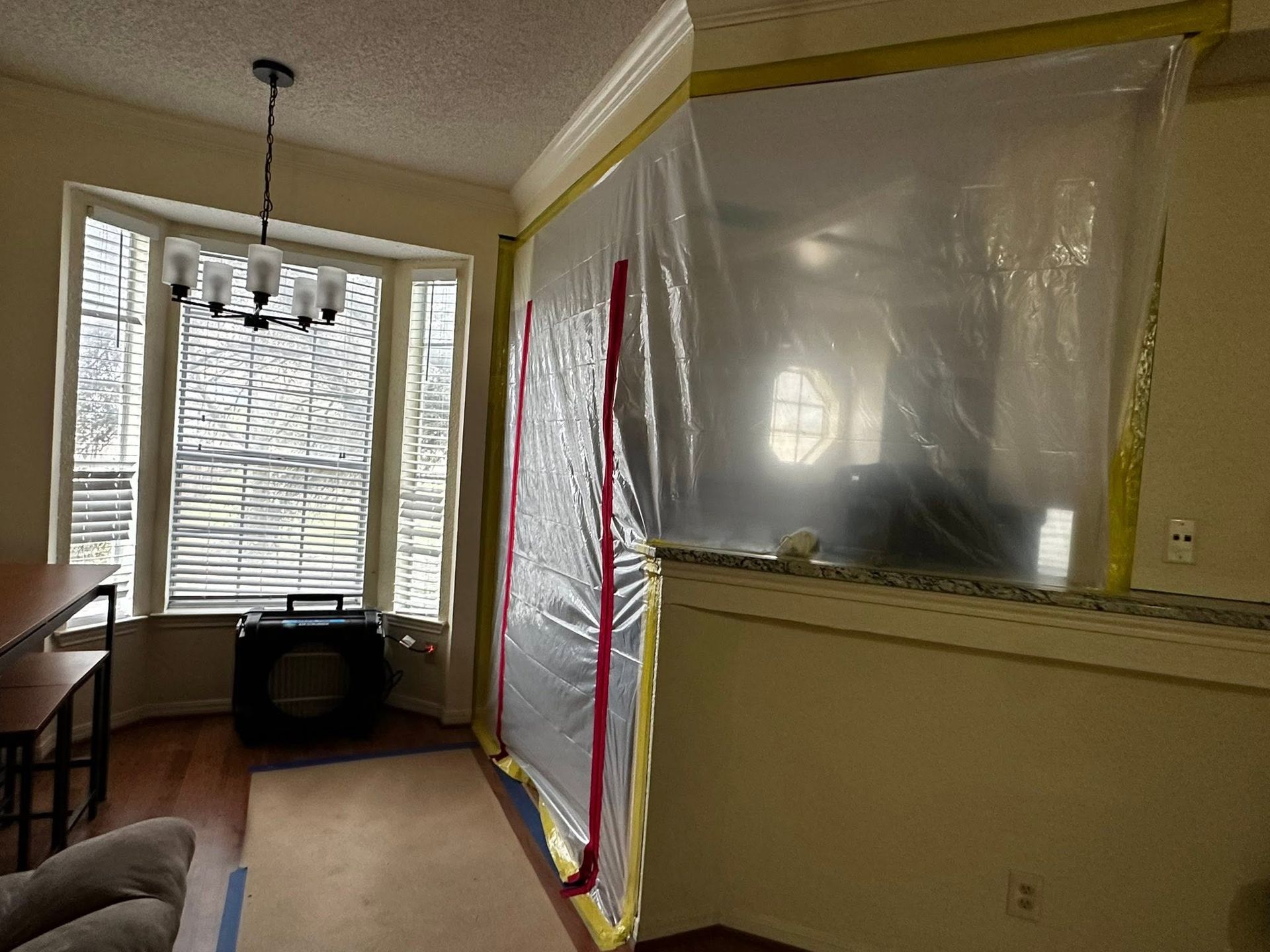 Living room with plastic-covered window, yellow trim, and blinds by a dining table and chair