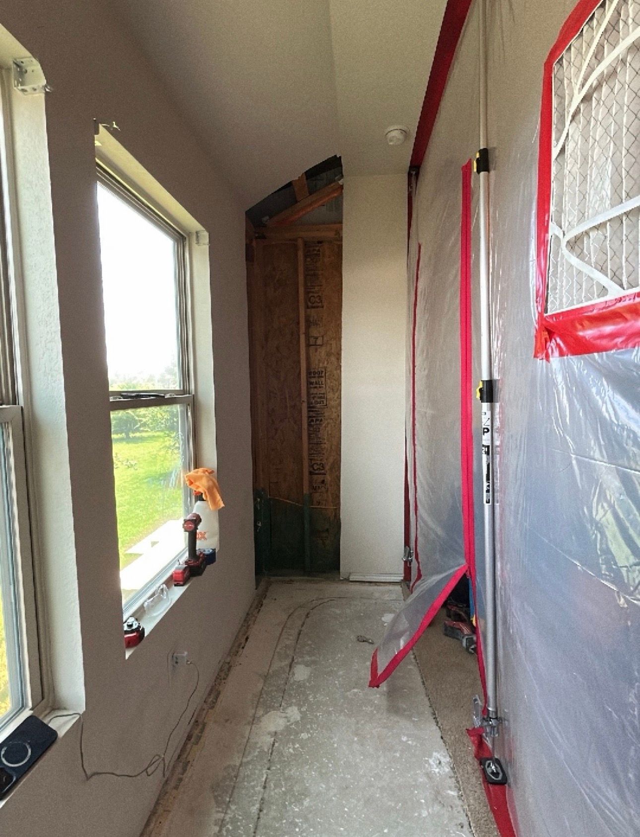 Narrow hallway under renovation with windows, plastic sheeting, and red taped masking on the right wall.