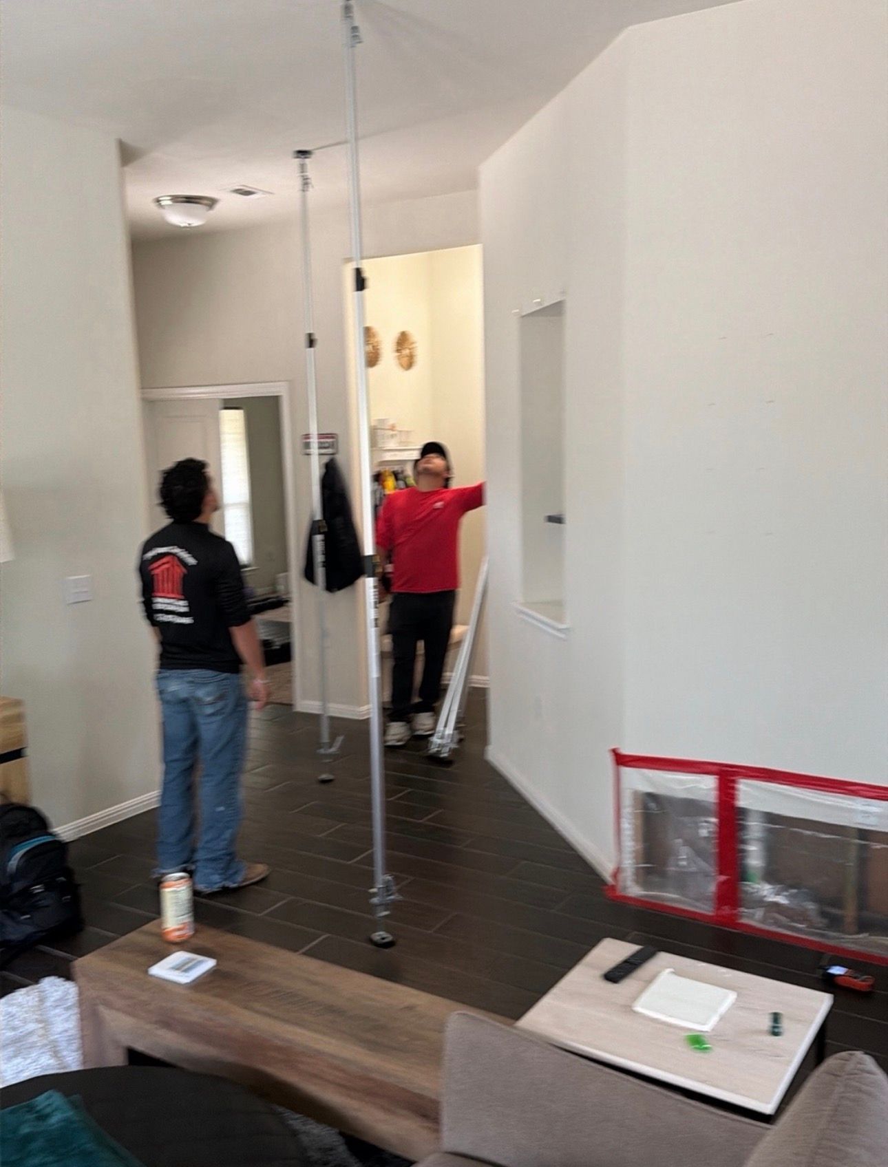 Two people in a white apartment room with dark wood floors, one standing by a doorway and one near a hanging pole.
