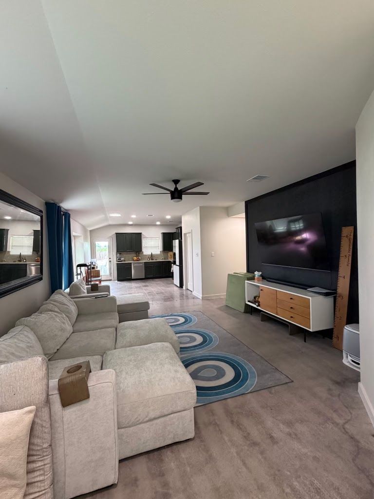 Modern open-concept living room with beige sectional, large TV, ceiling fan, and kitchen in the background
