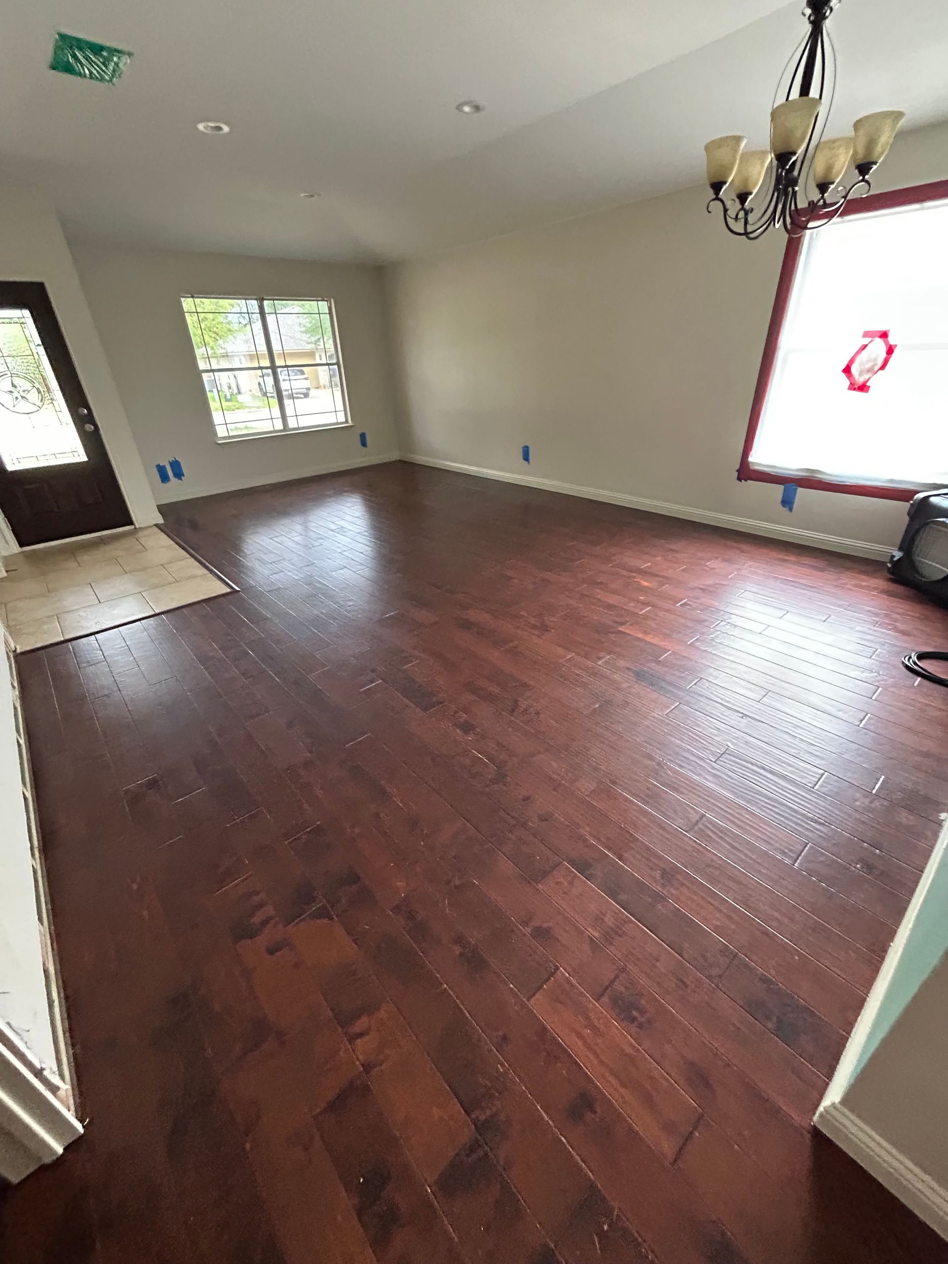 Empty living room with dark hardwood floors, white walls, chandelier, and large windows.