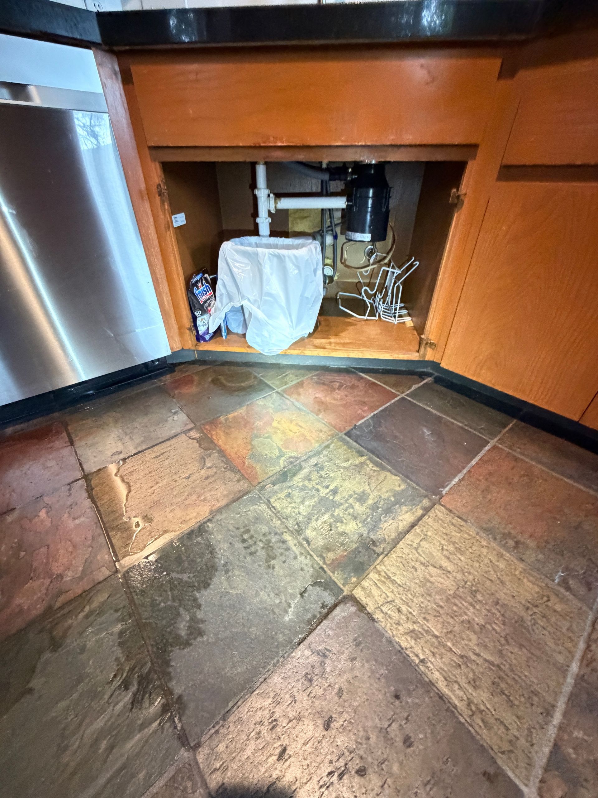 Kitchen sink cabinet with exposed plumbing and a cloth-covered item, on a tiled floor.