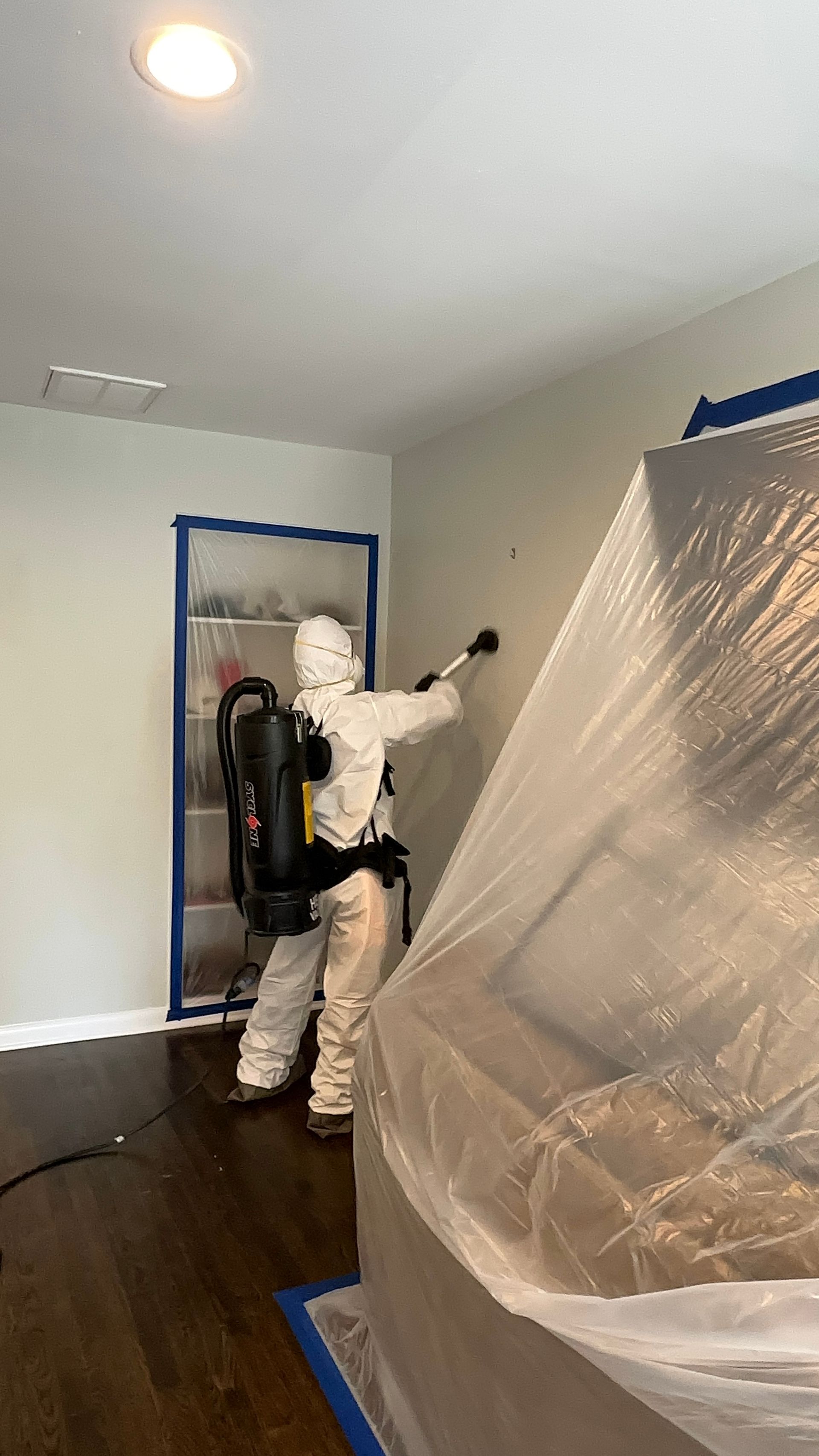 Person in a white suit sprays a wall in a room under renovation, with furniture covered in plastic.
