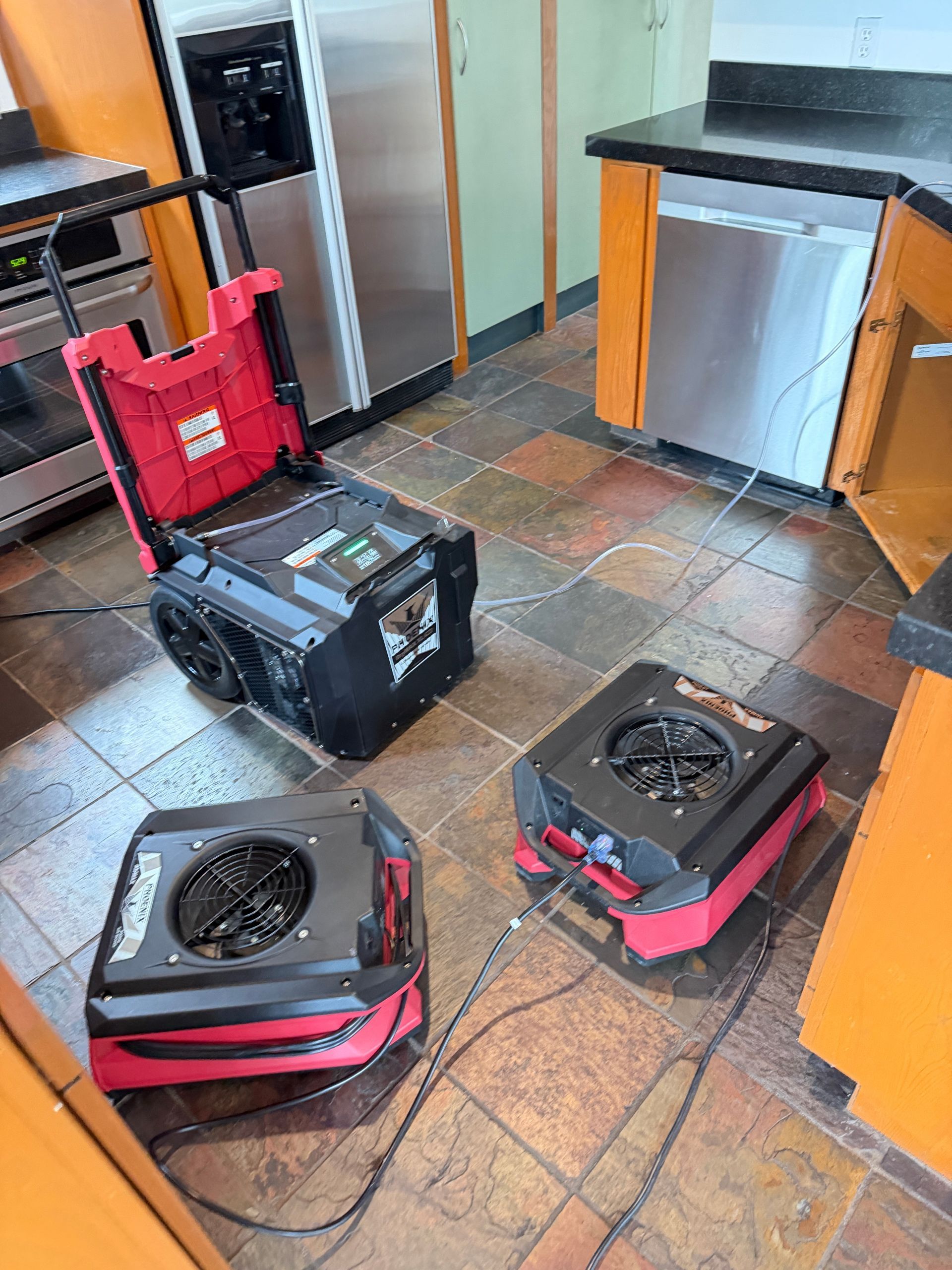Two portable cooling units and a black equipment case on a tiled kitchen floor with stainless steel appliances.
