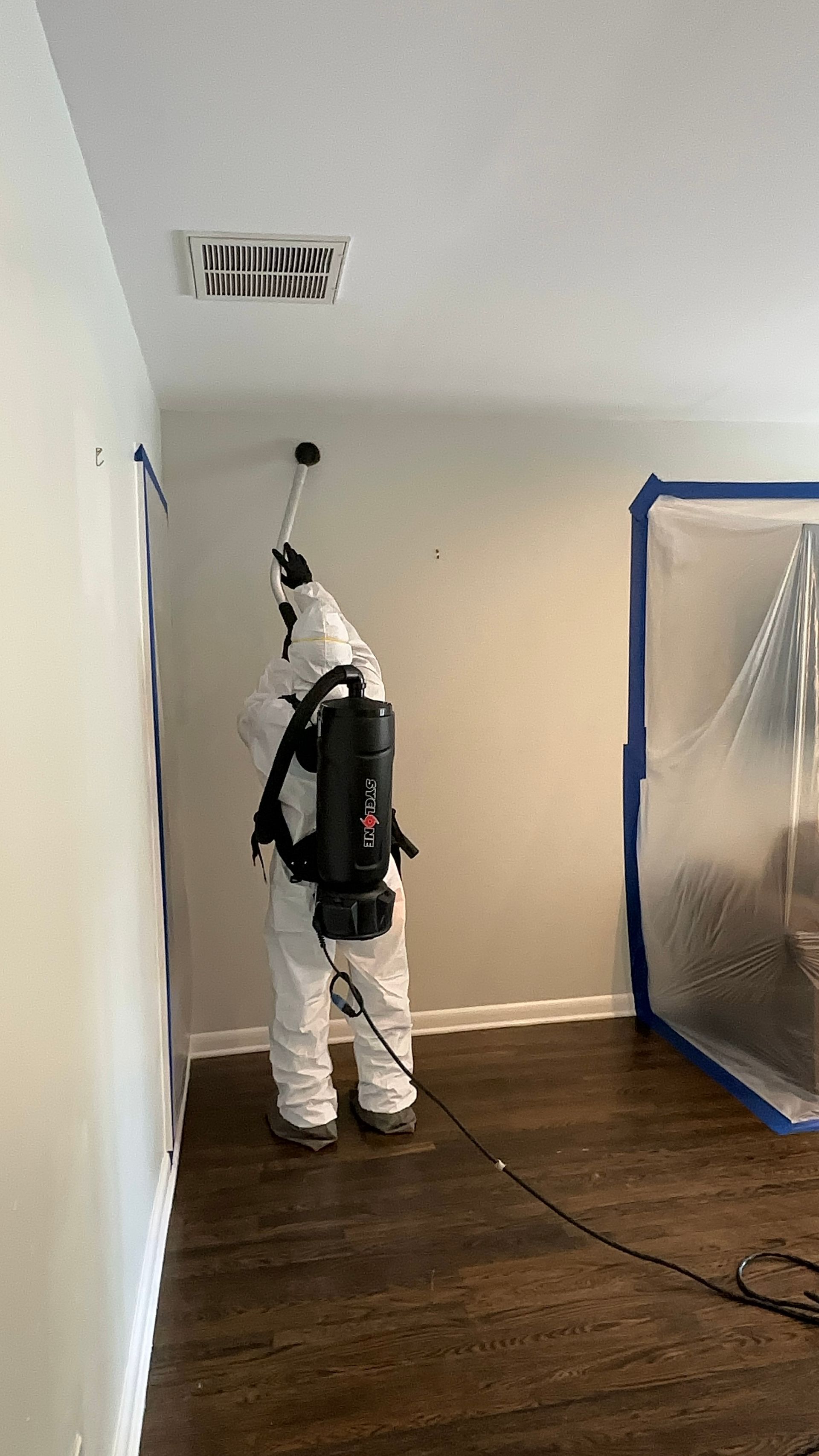 Person in white protective suit spraying or cleaning a wall inside a room, with a plastic-covered area nearby.