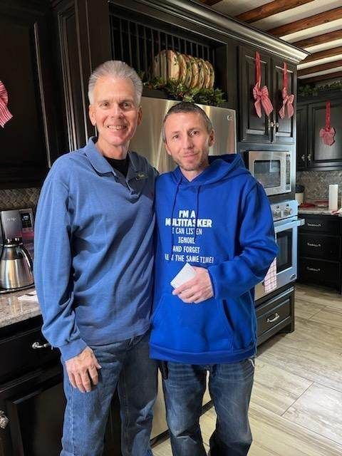 Two men are posing for a picture in a kitchen . one of the men is wearing a blue hoodie.