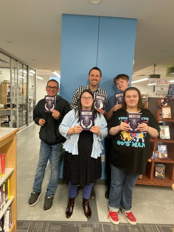 Four people holding books stand in a library aisle, smiling in front of blue cabinets.