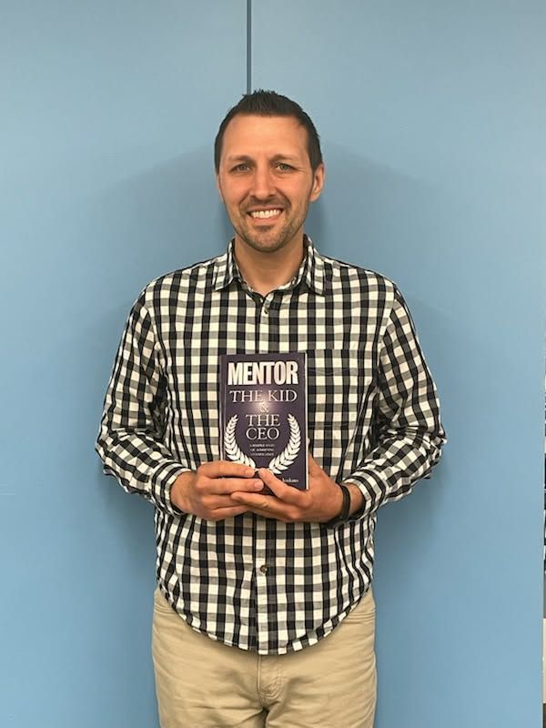Man standing against a blue wall, smiling and holding a book with a dark purple cover.