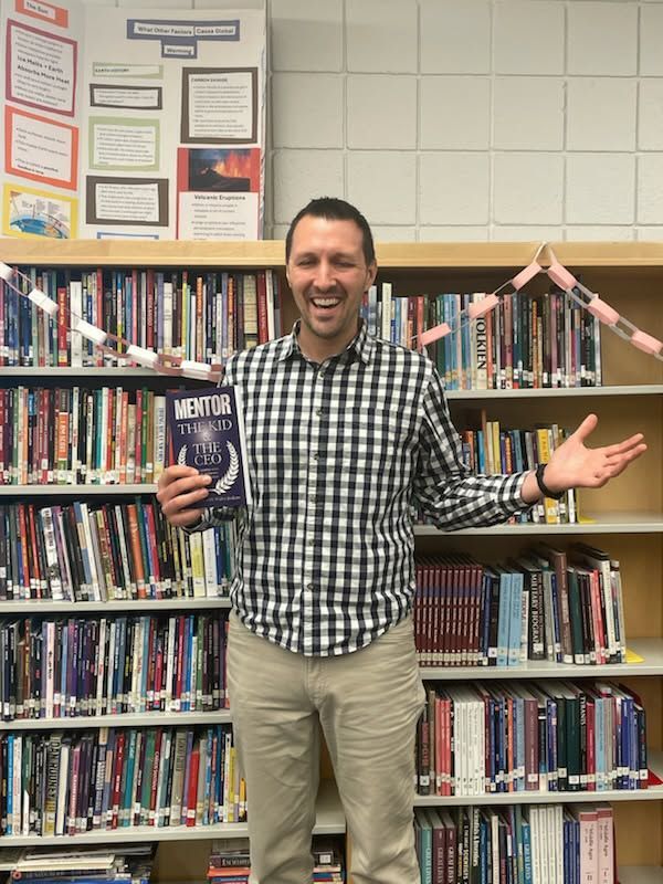Smiling person holding a book in a library aisle, standing in front of shelves and raising both arms.