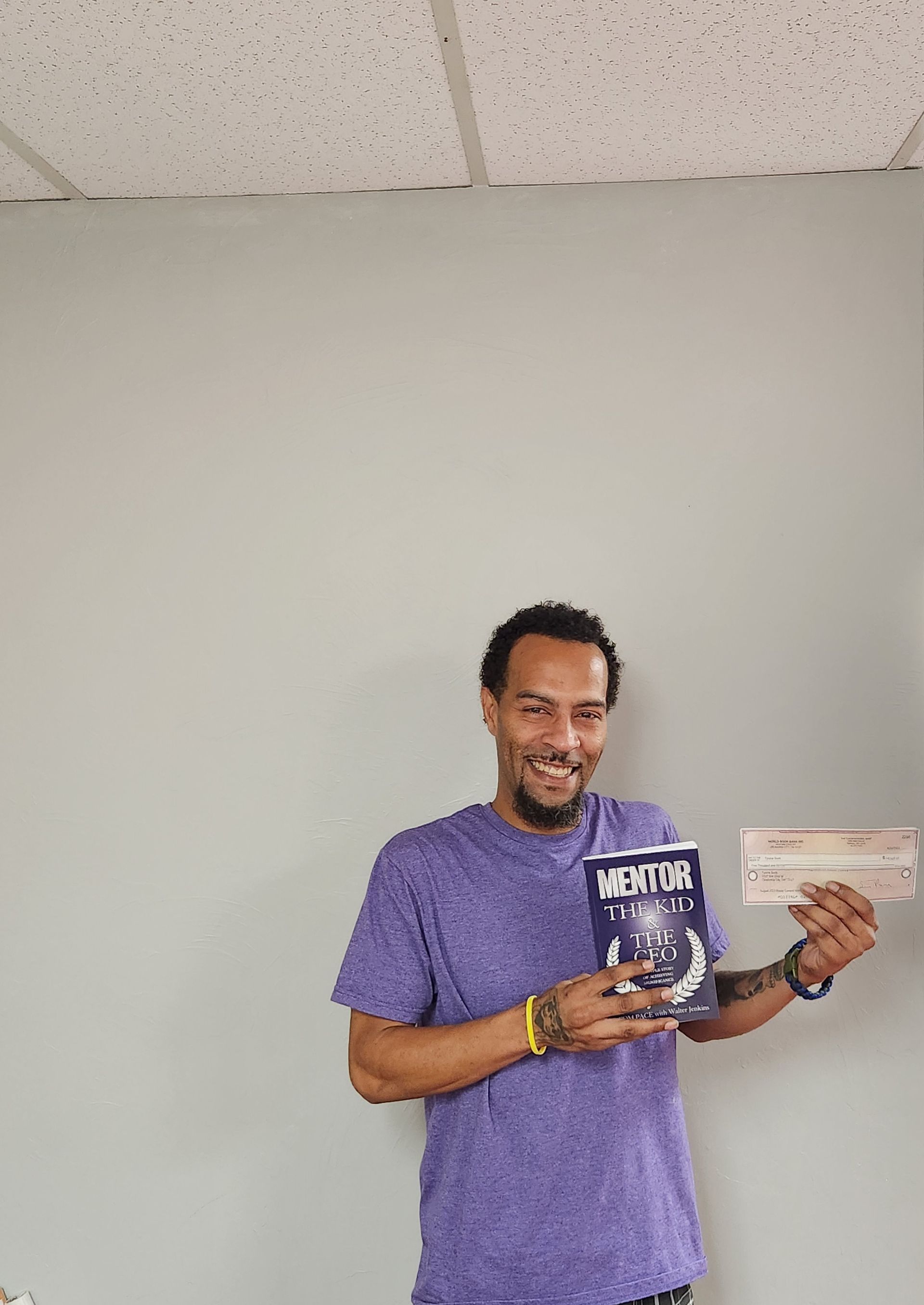 A man in a purple shirt is holding a book and a check.