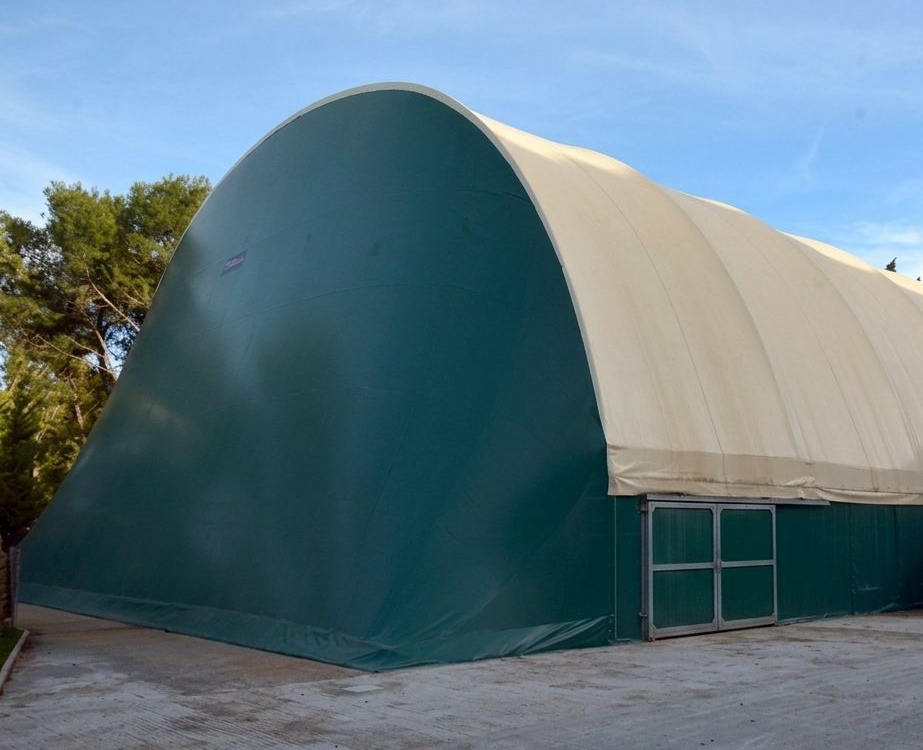 Grande tenda da deposito ad arco verde e beige su un piazzale pavimentato sotto un cielo azzurro
