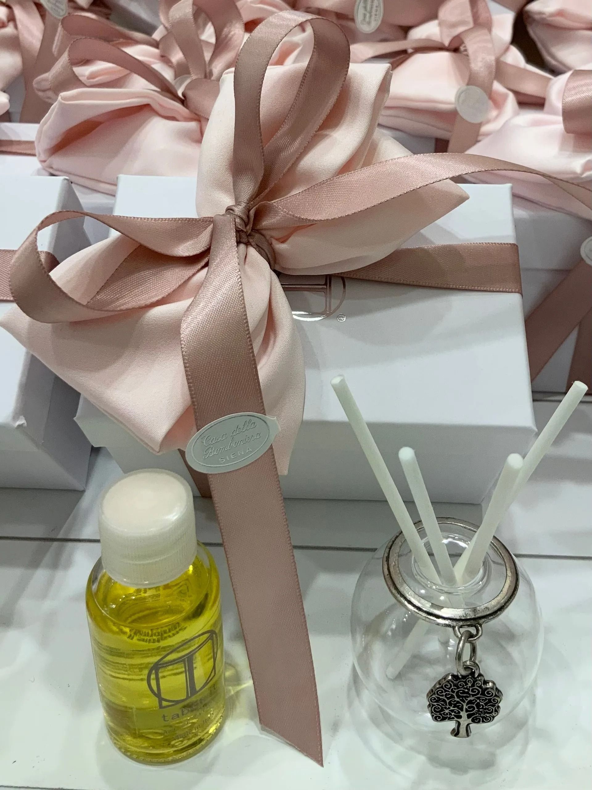 Confezione regalo con nastro rosa, bottiglietta gialla e diffusore a bastoncini su una superficie chiara.