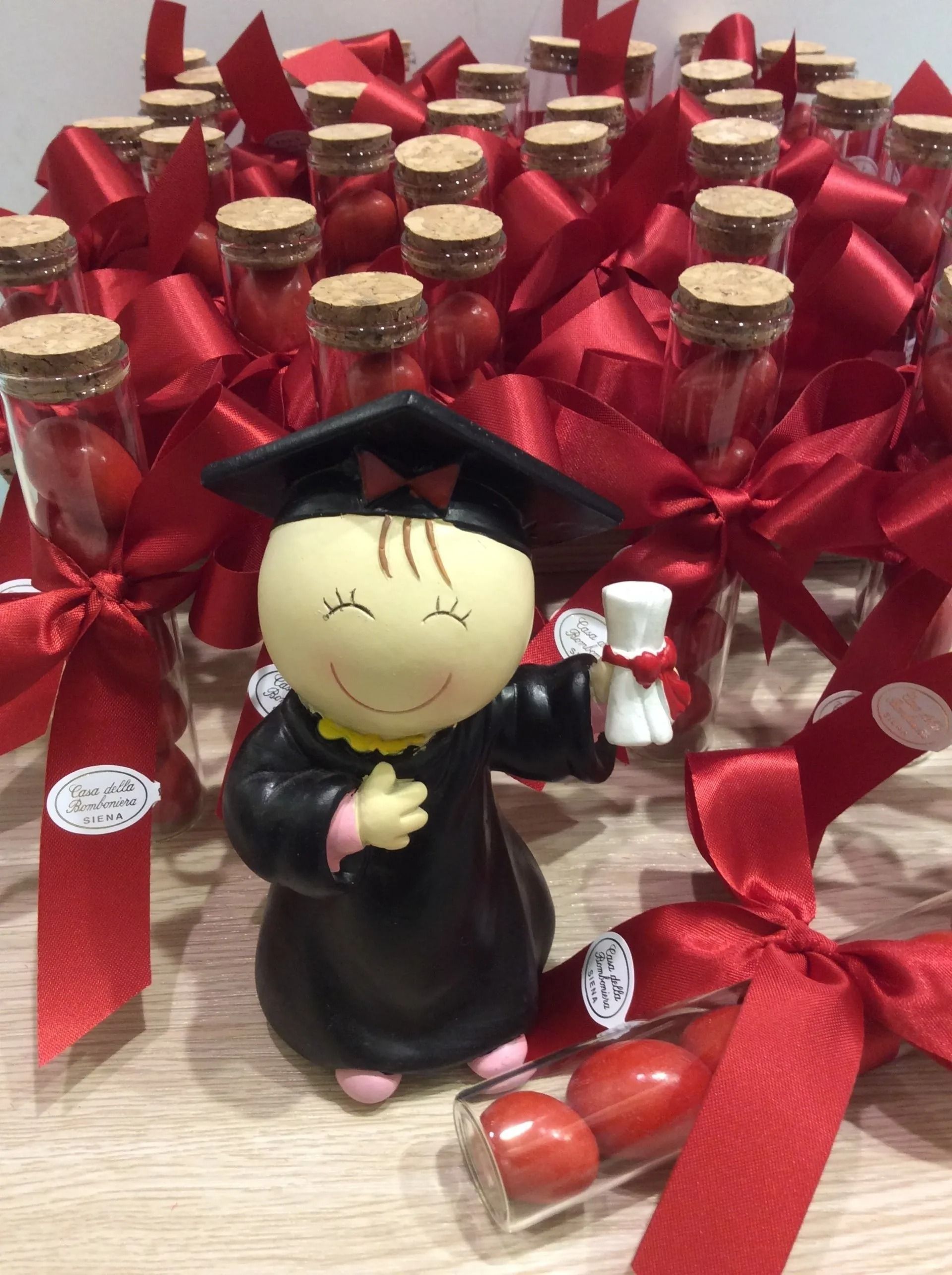 Bambola di laurea che tiene in mano un diploma, circondata da bottiglie avvolte in nastri rossi e bomboniere.