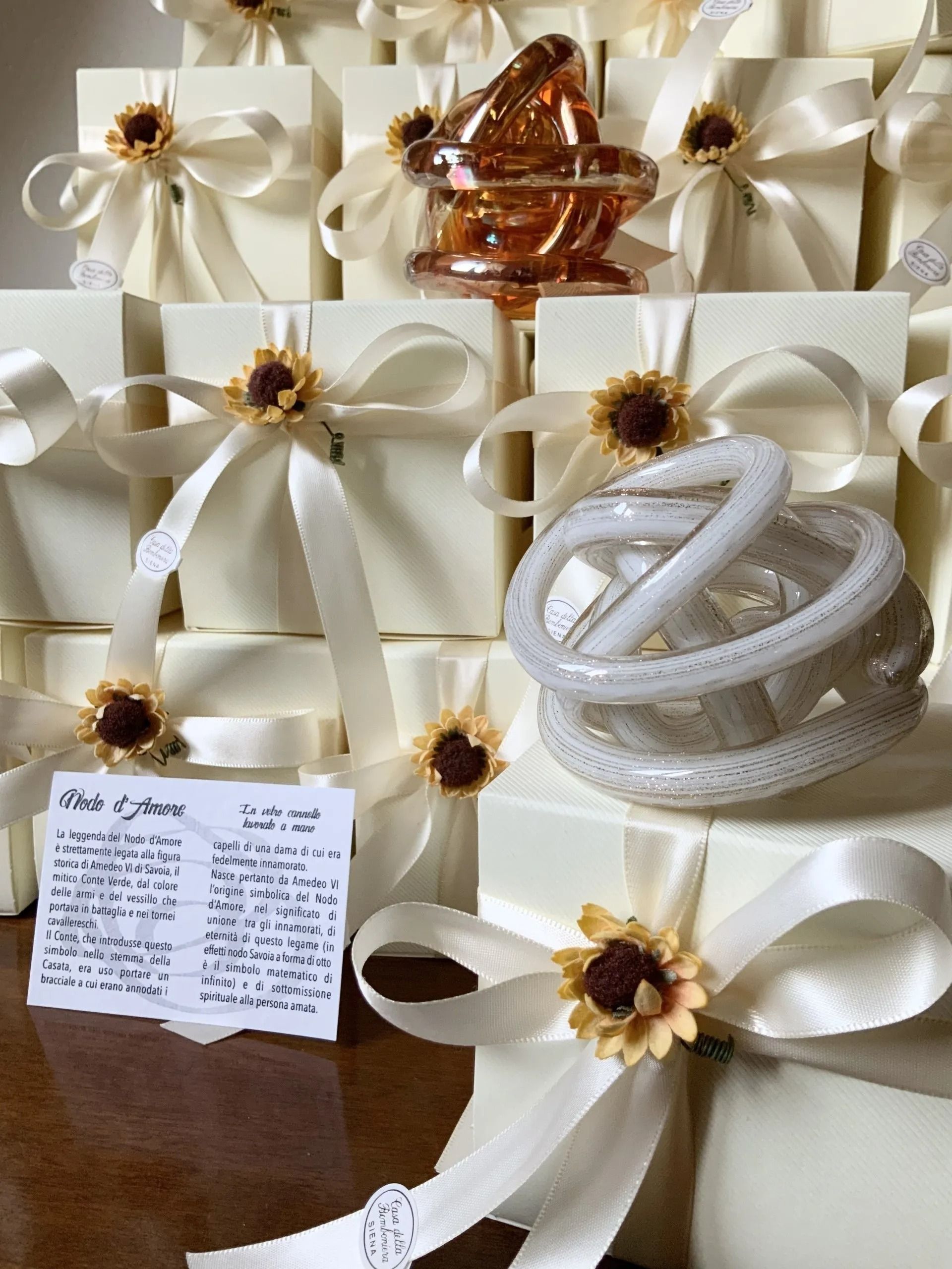 Scatole regalo color crema legate con nastri bianchi e decorazioni a forma di girasole, con cordoncino bianco arrotolato e un elemento decorativo color ambra.