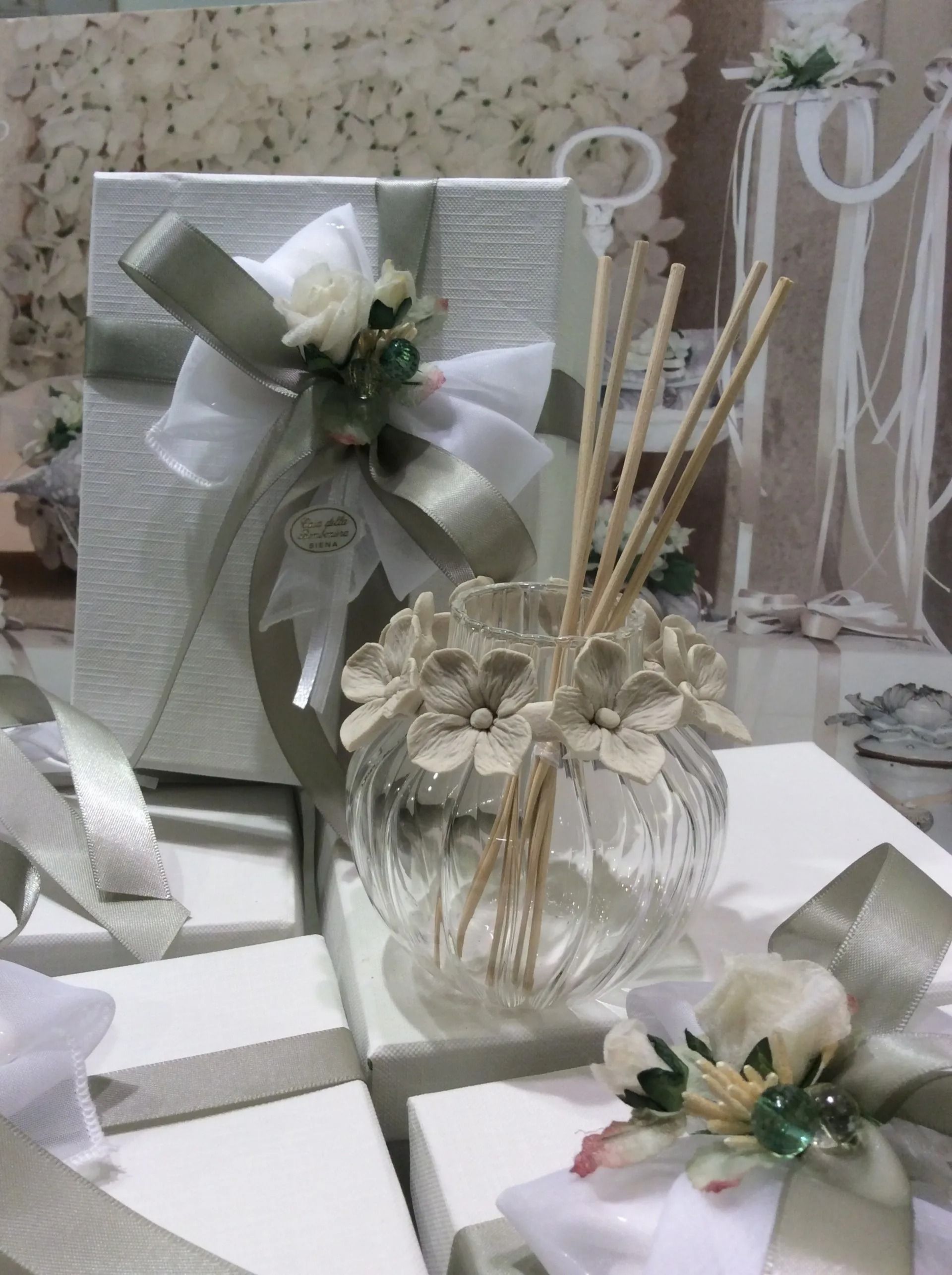 Sacchetti regalo bianchi e bomboniere floreali legate con un nastro argentato, disposte su un tavolo con bastoncini decorativi e un tocco di pizzo.