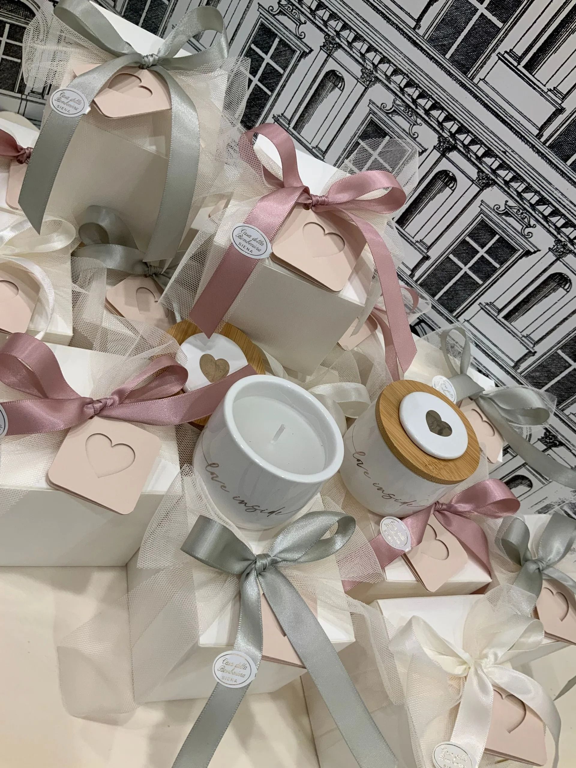 Scatole regalo dai toni pastello con nastri rosa, una candela bianca e un piccolo barattolo, il tutto disposto su carta da regalo decorata.