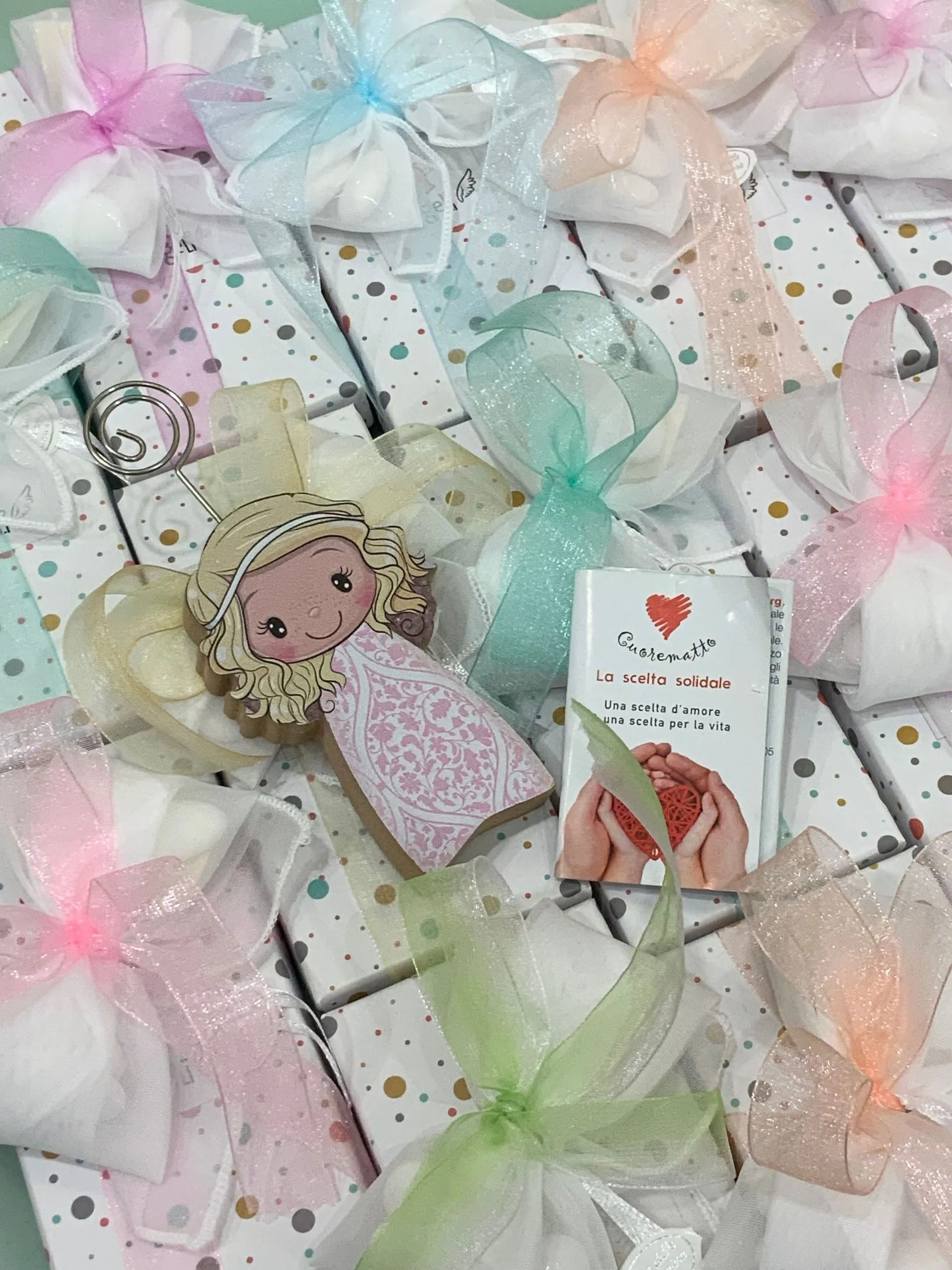 Sacchetti regalo color pastello e un piccolo biglietto a forma di bambola su una superficie piastrellata con puntini simili a coriandoli.