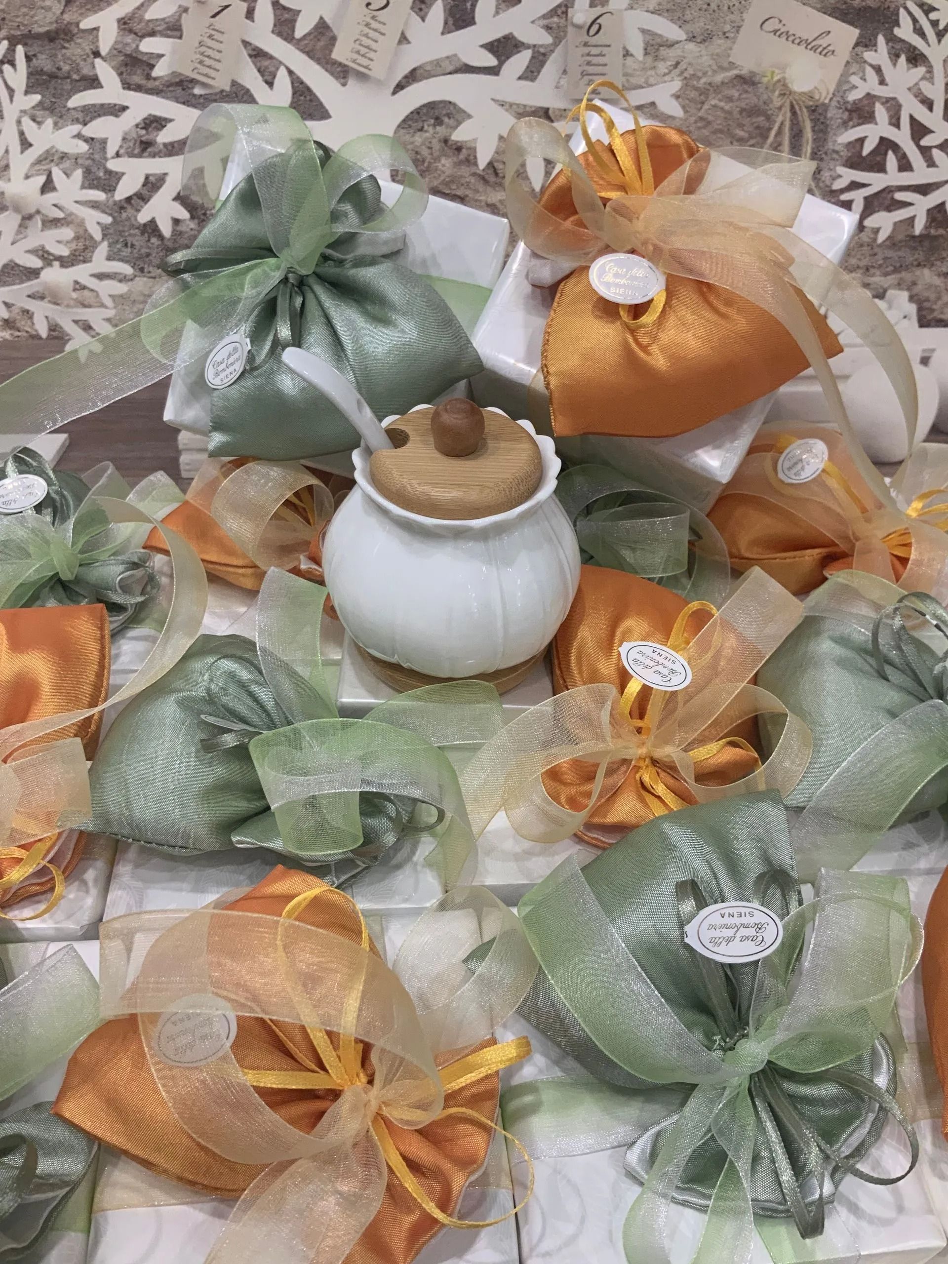 Sacchetti regalo in organza assortiti, verdi e arancioni, disposti intorno a un piccolo barattolo bianco su una superficie decorata.