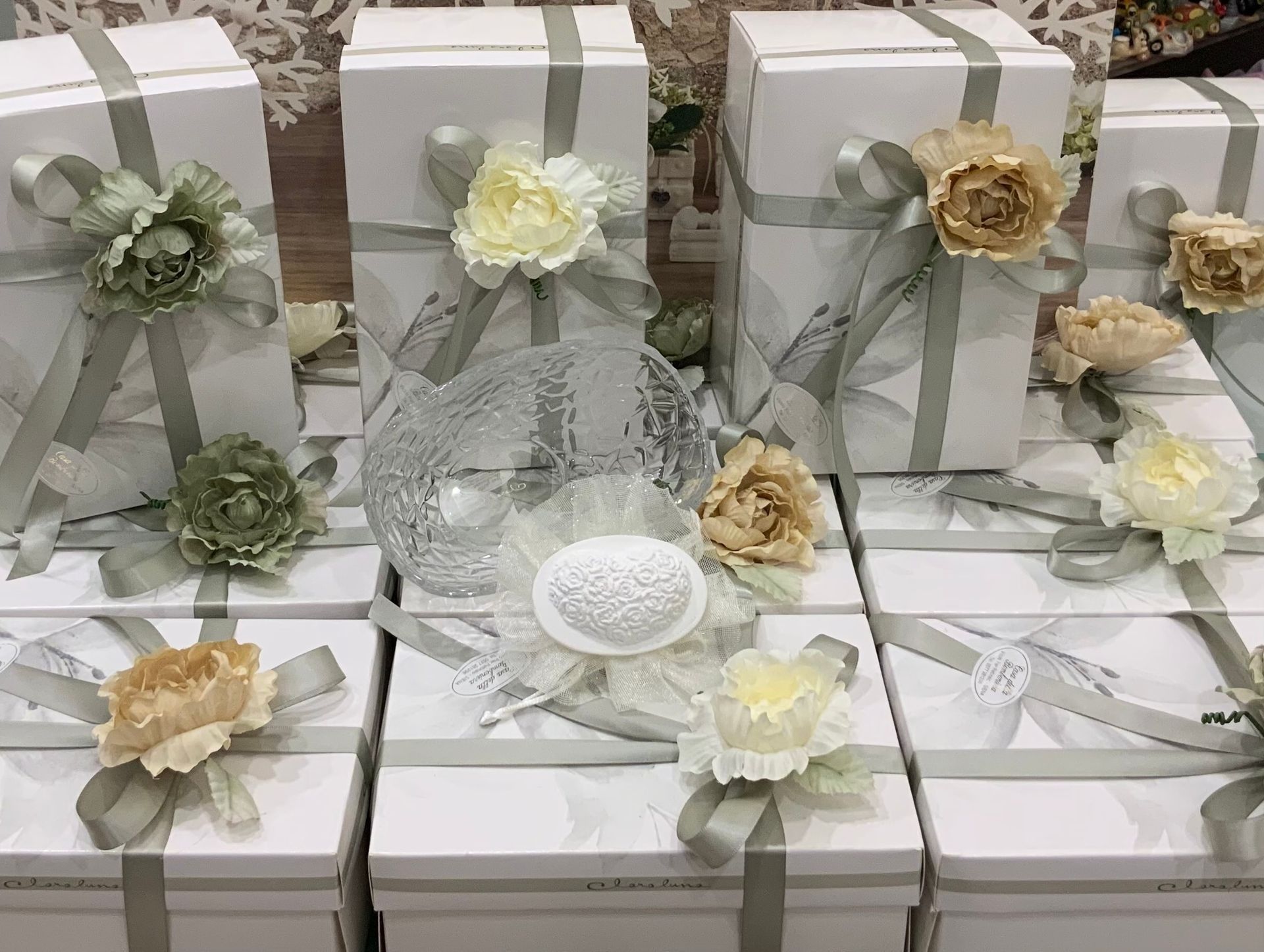 Scatole regalo bianche con nastro e fiori dai colori pastello disposti intorno a una ciotola di cristallo su un tavolo