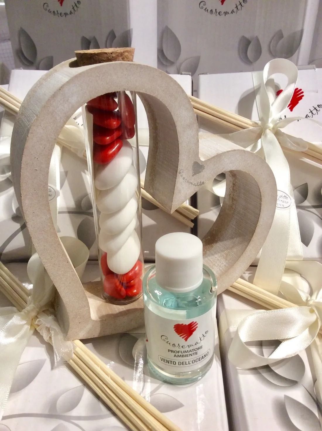Composizione decorativa a forma di cuore con candela rossa, diffusori a bastoncini e una bottiglietta su una superficie decorata.