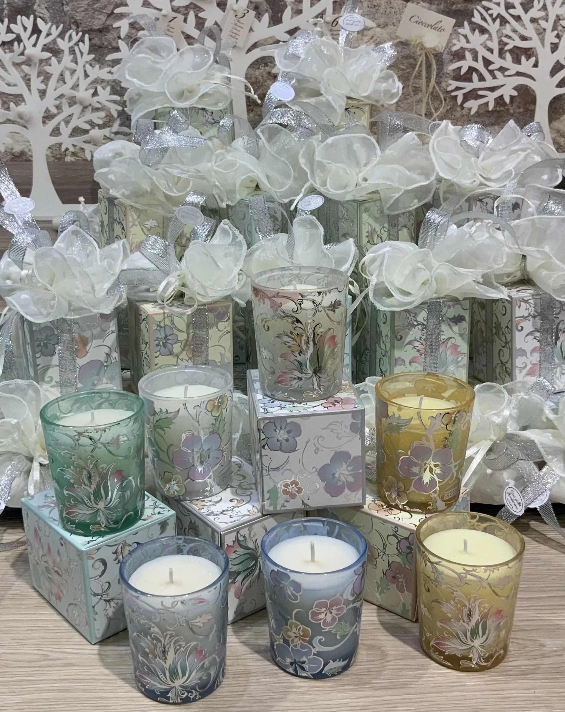 Vasi portacandele con decorazioni floreali pastello e fiocchi bianchi, disposti su più livelli su una superficie di legno.