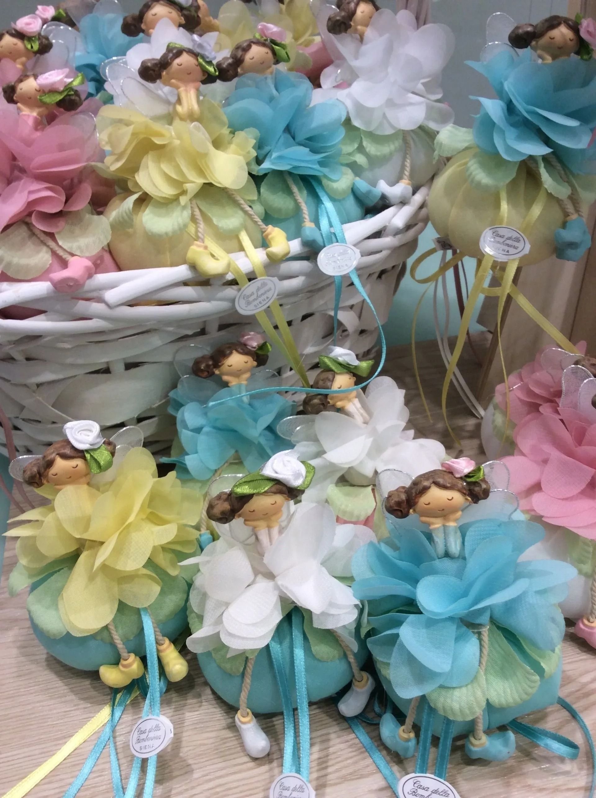 Assortimento di bambole floreali dai colori pastello con ali di pizzo in un cestino di vimini, nei colori giallo, blu, rosa e bianco.