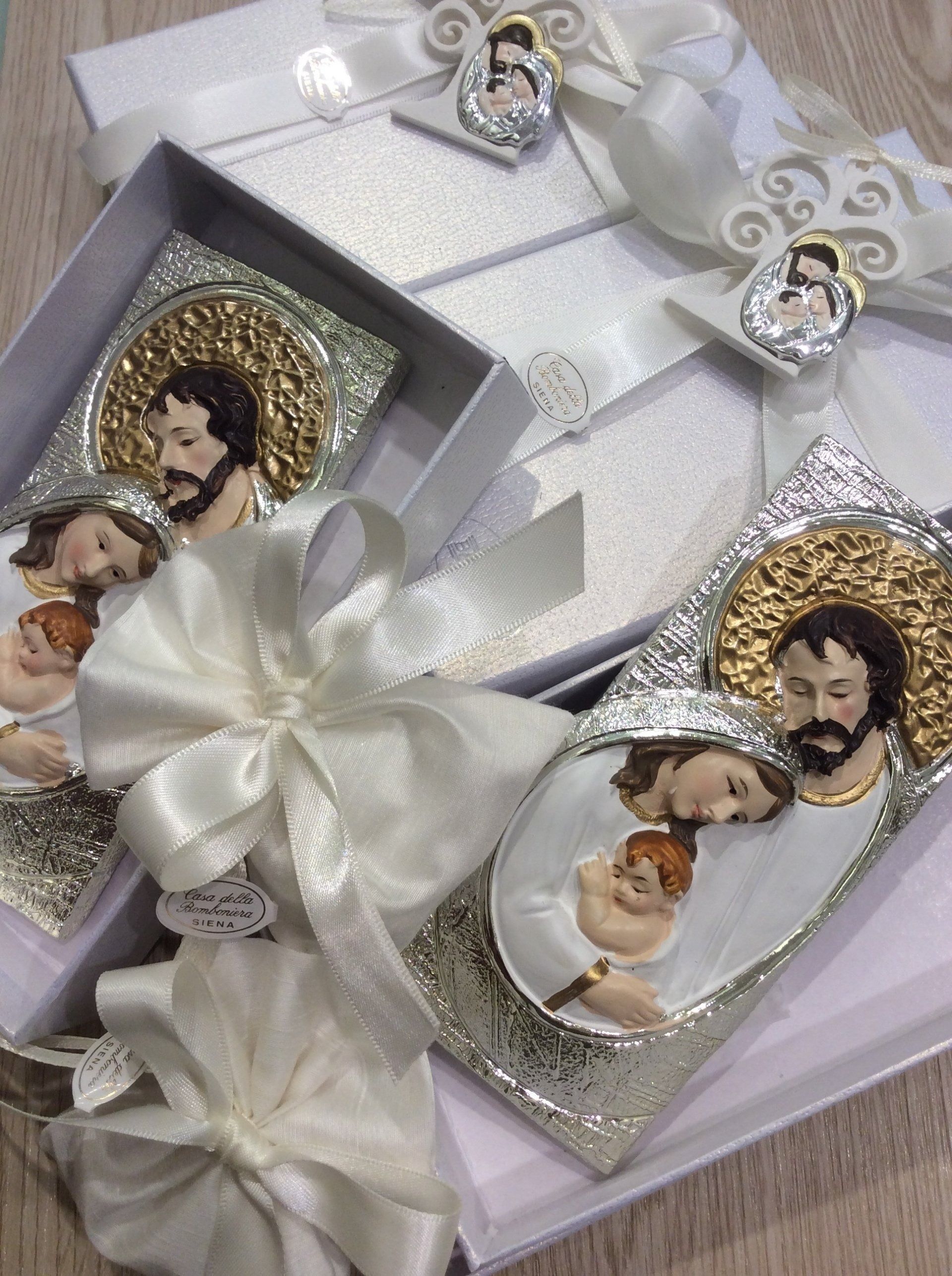 Ornamenti religiosi in argento con piccoli ritratti sacri e fiocchi di raso bianco su uno sfondo di tessuto chiaro.