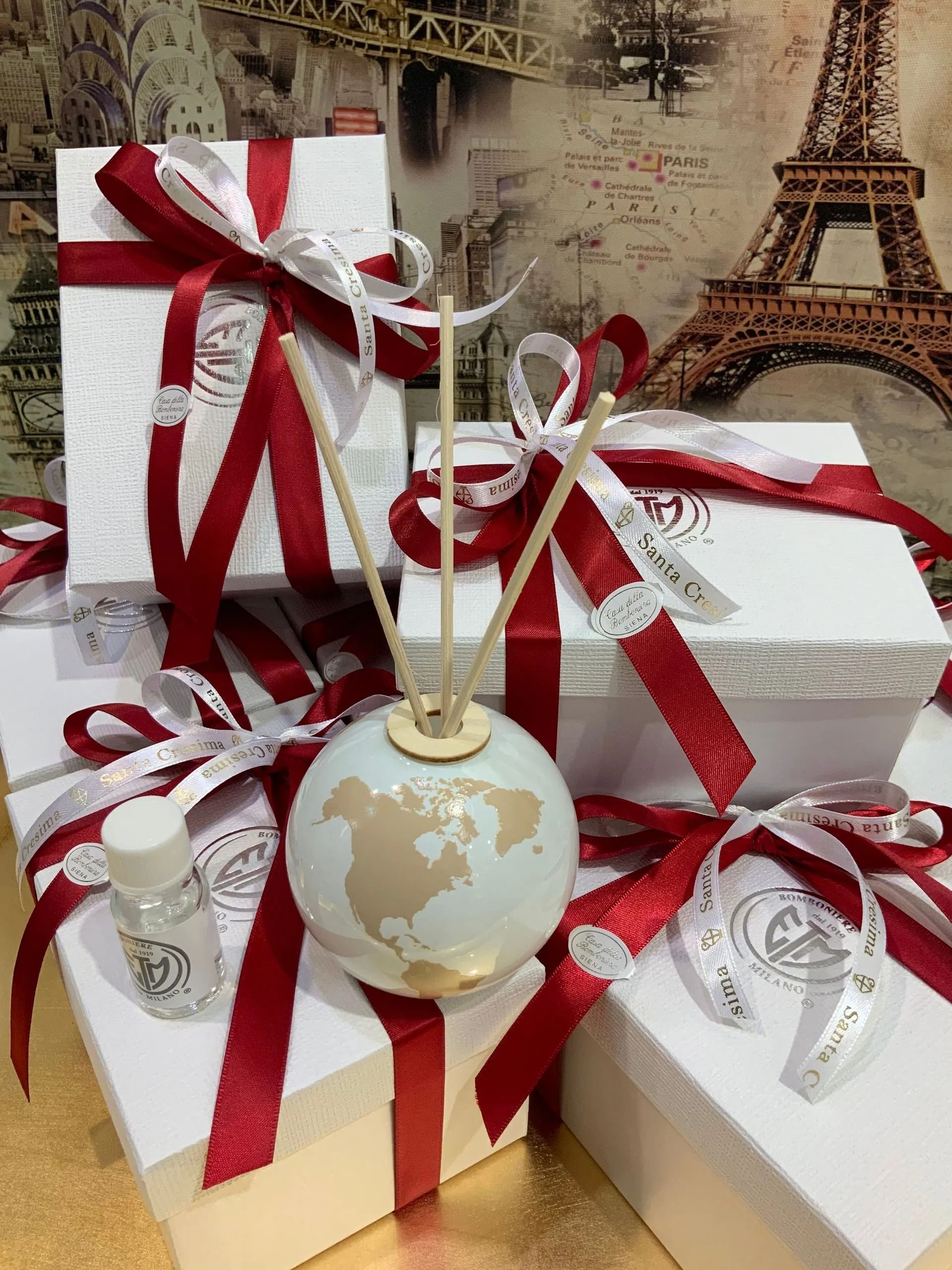 Scatole regalo natalizie legate con nastri rossi e un diffusore bianco-oro davanti a uno sfondo raffigurante la Torre Eiffel.