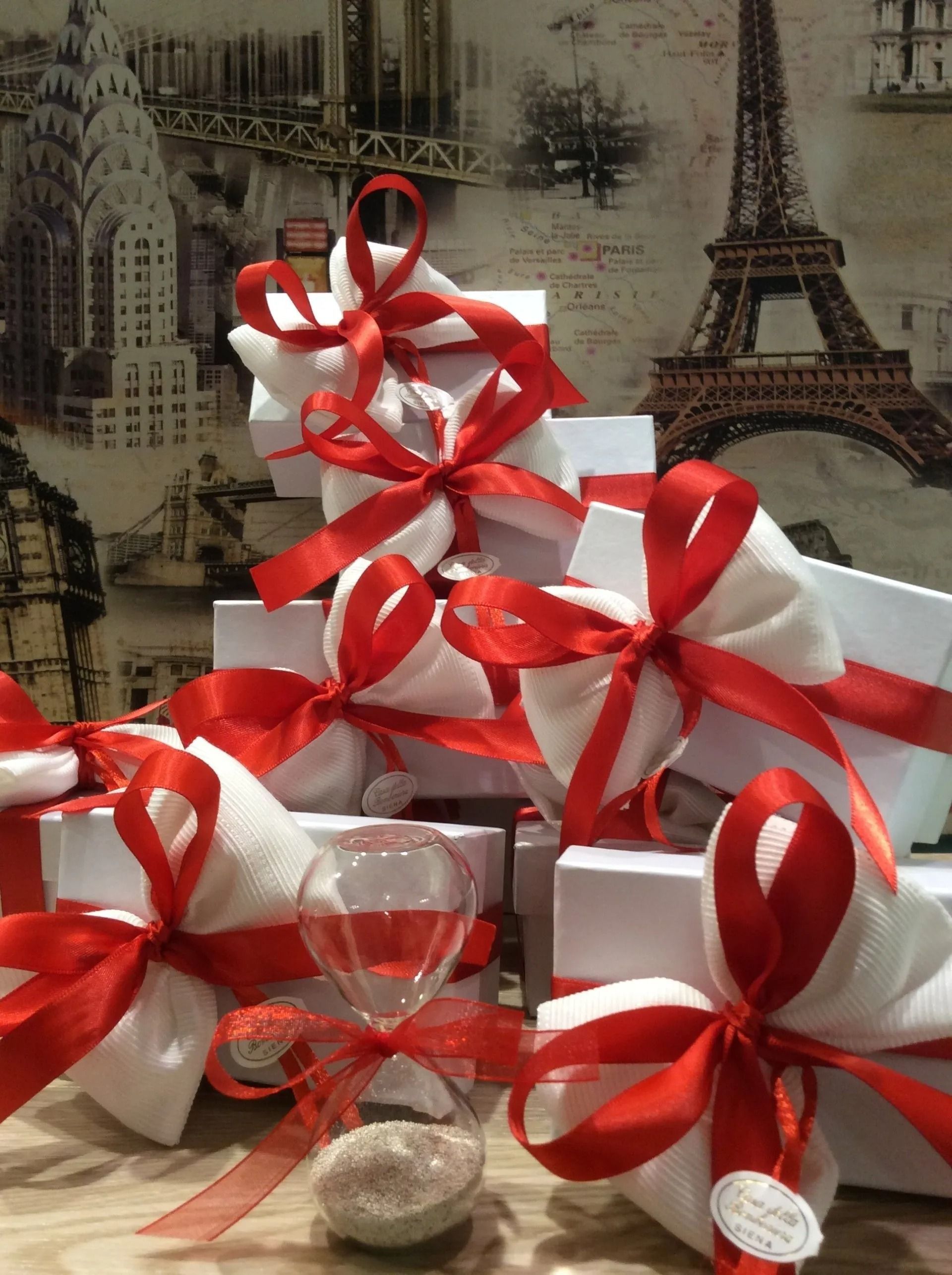 Scatole regalo decorate con fiocchi di nastro bianco e nastri di raso rosso, disposte davanti a uno sfondo raffigurante la Torre Eiffel.