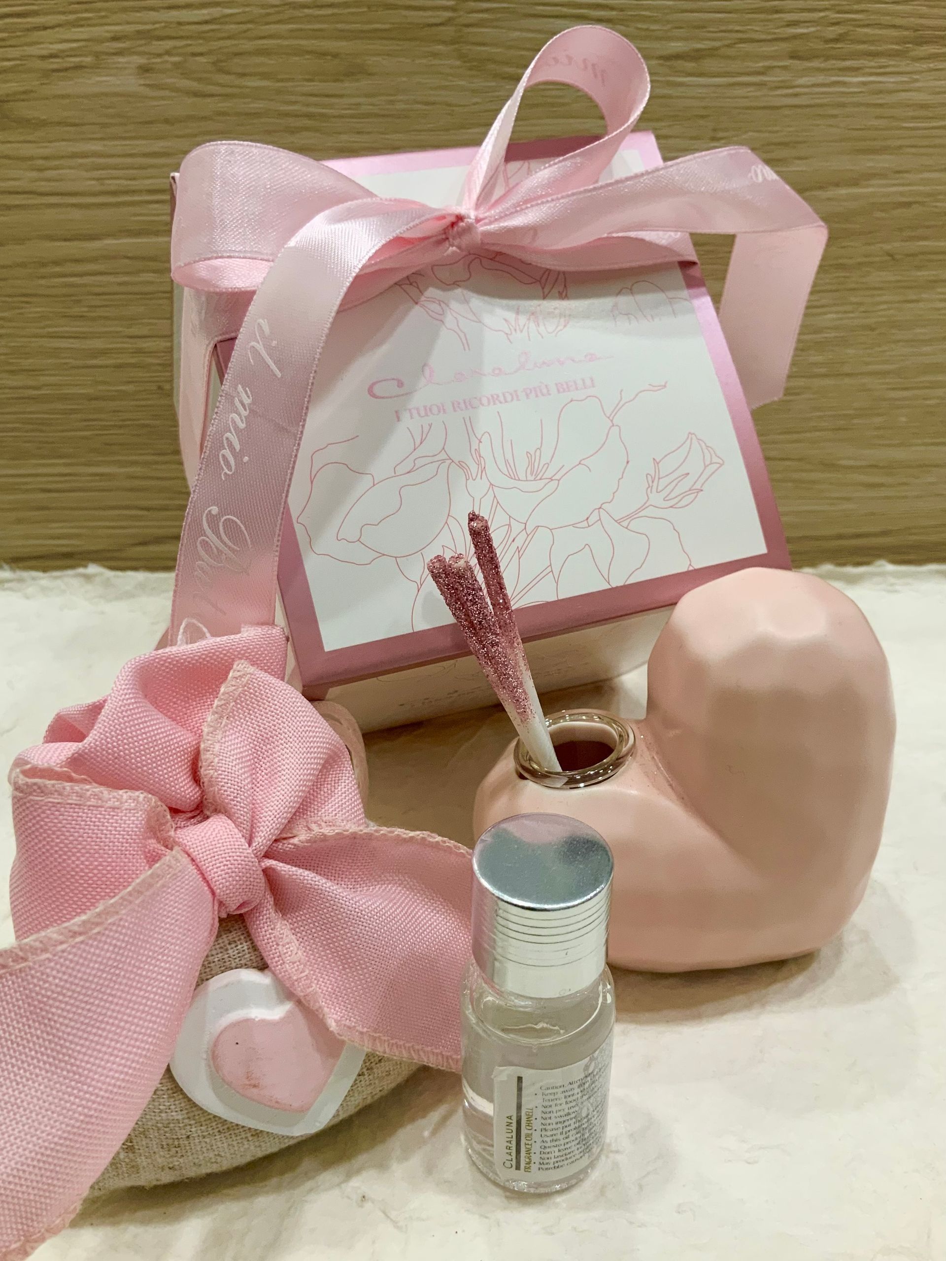 Scatola regalo rosa con nastro, fiocco, candela e bottiglietta su una superficie beige.