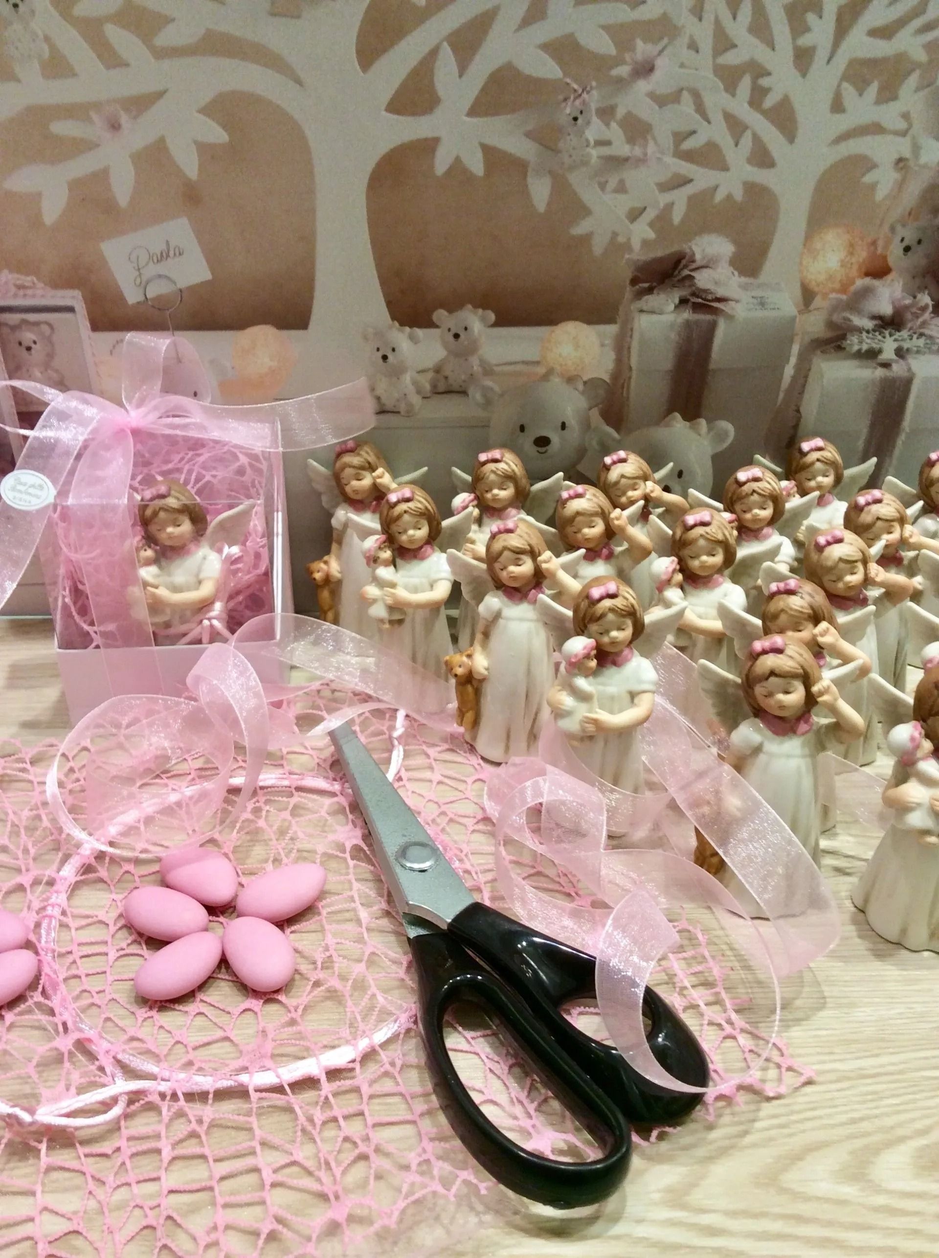 Esposizione di lavoretti creativi rosa con pizzo, forbici, nastro e tante piccole statuette di angeli disposte su un tavolo