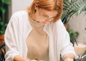 Woman with red hair, wearing white shirt, writing at a table in a cafe, drink in front of her.