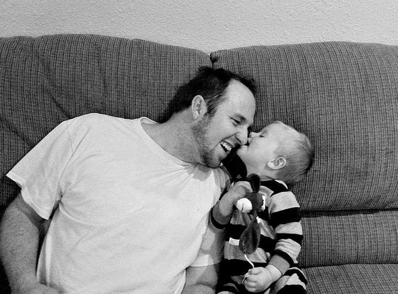 A parent smiles while sitting on a couch, gently touching noses with a toddler who is holding a small toy.