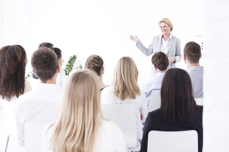 A professional presenting to an audience in a bright, modern classroom or conference room setting.
