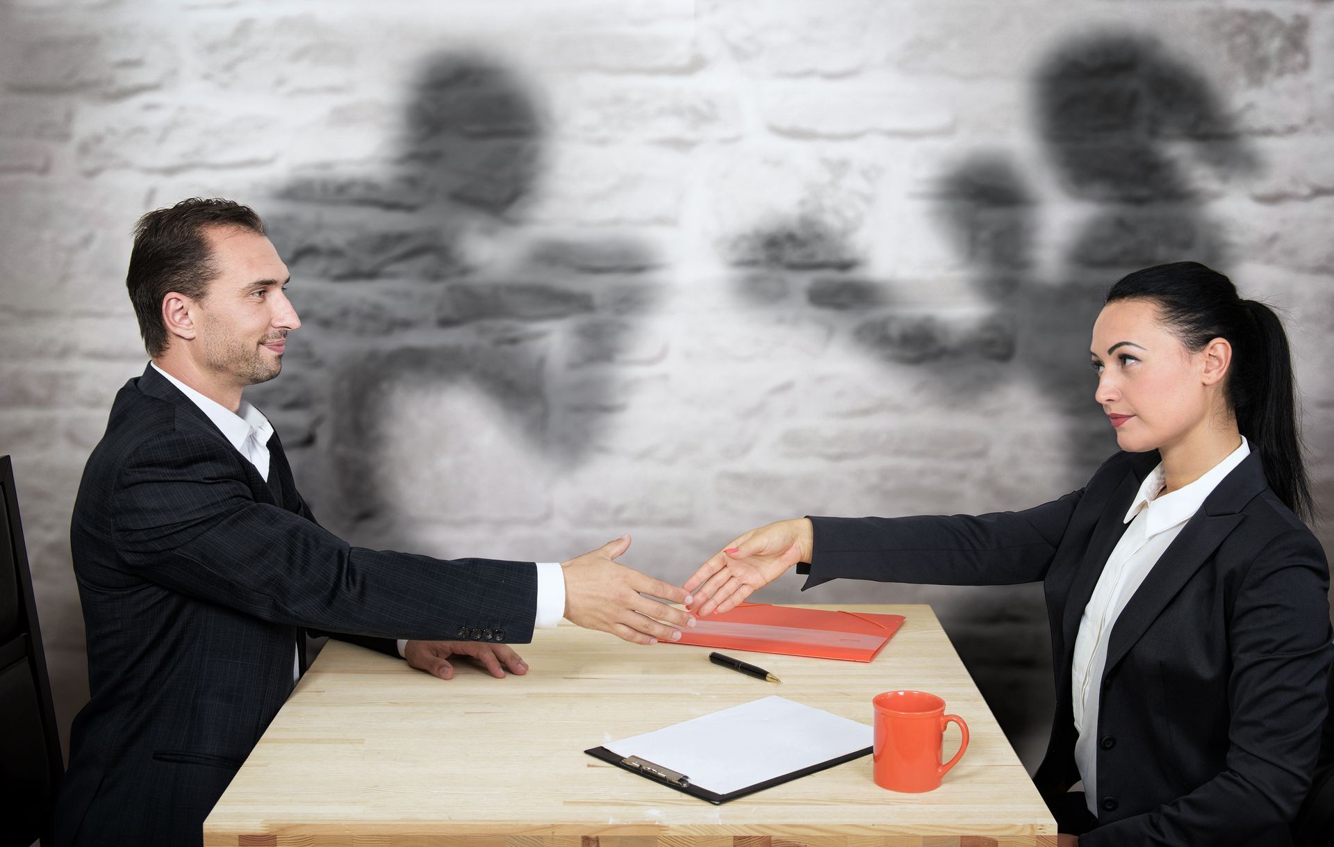 Two people in business attire reach to shake hands across a desk, with shadowy figures of boxers on the wall behind them.